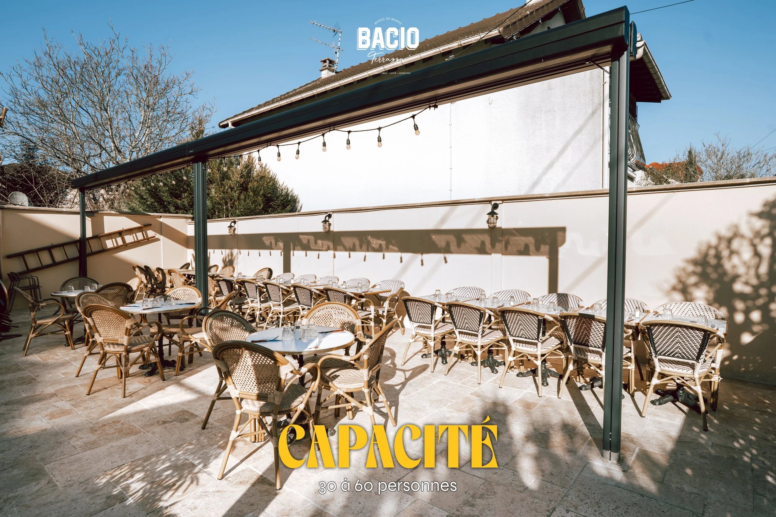 Terrasse extérieure avec plusieurs tables et chaises en rotin, entourée de murs blancs, sous un toit avec un éclairage suspendu, dans un cadre ensoleillé et ombragé, avec des arbres dénudés en arrière-plan, annonçant un restaurant ou café nommé 'Bacio' avec capacité de 30 à 60 personnes.
