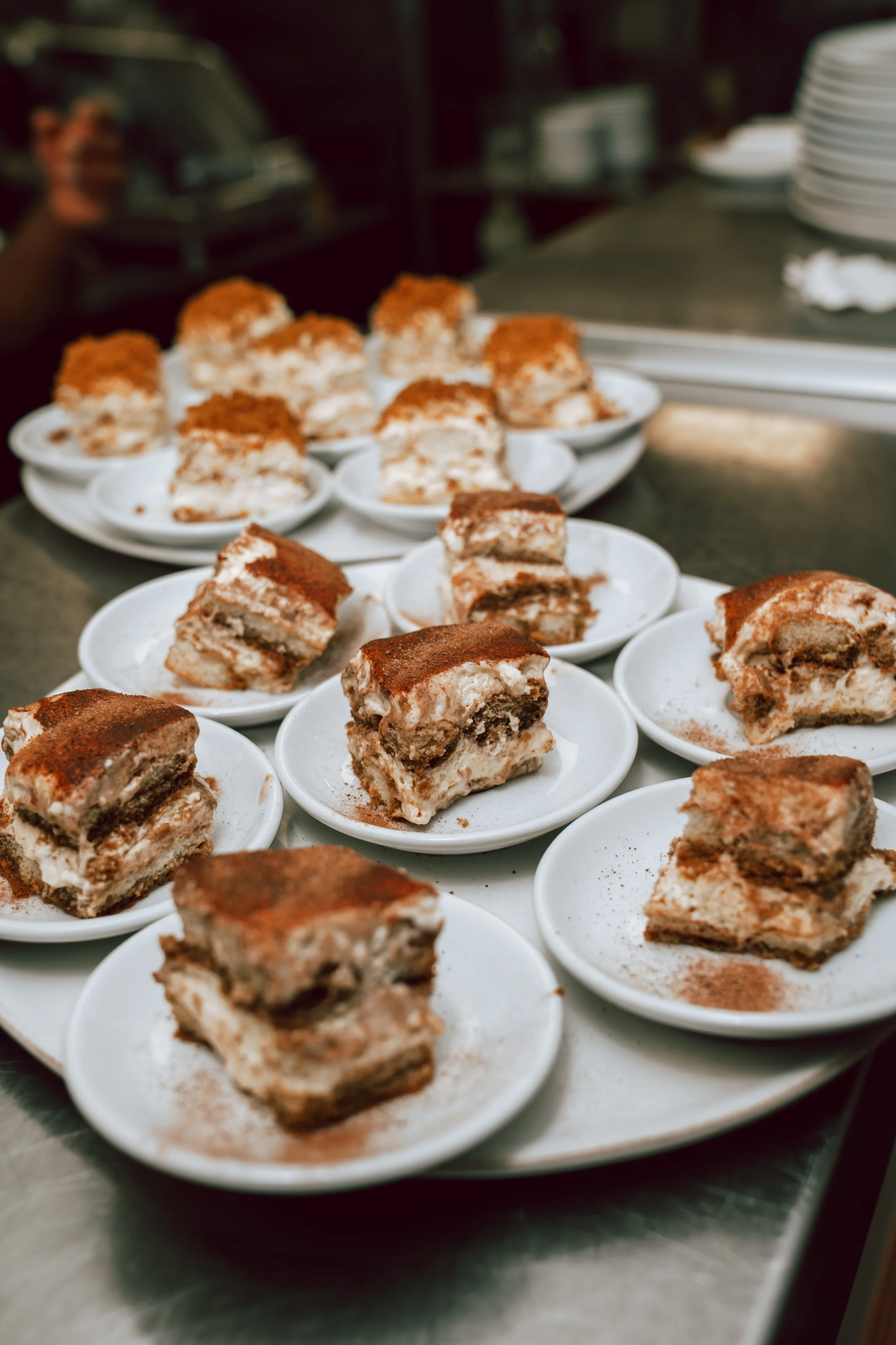 Gros plan de plusieurs parts de tiramisu sur de petites assiettes blanches, disposées sur une grande plaque dans une cuisine.