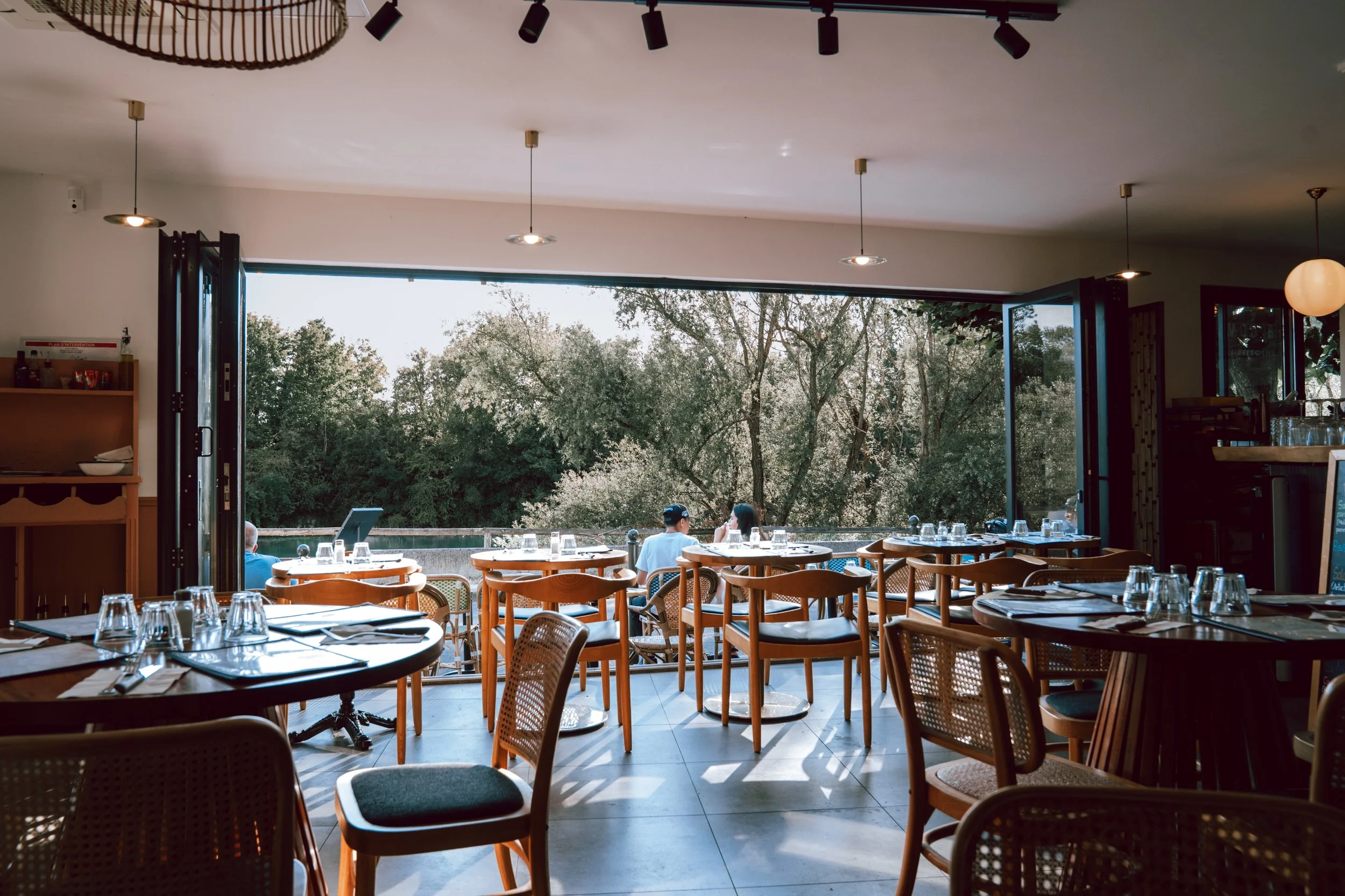 Intérieur d'un restaurant avec des tables en bois, des chaises en rotin, une grande baie vitrée ouverte donnant sur un espace vert, et des lampes suspendues. Des gens sont assis, en train de parler et de profiter du cadre.