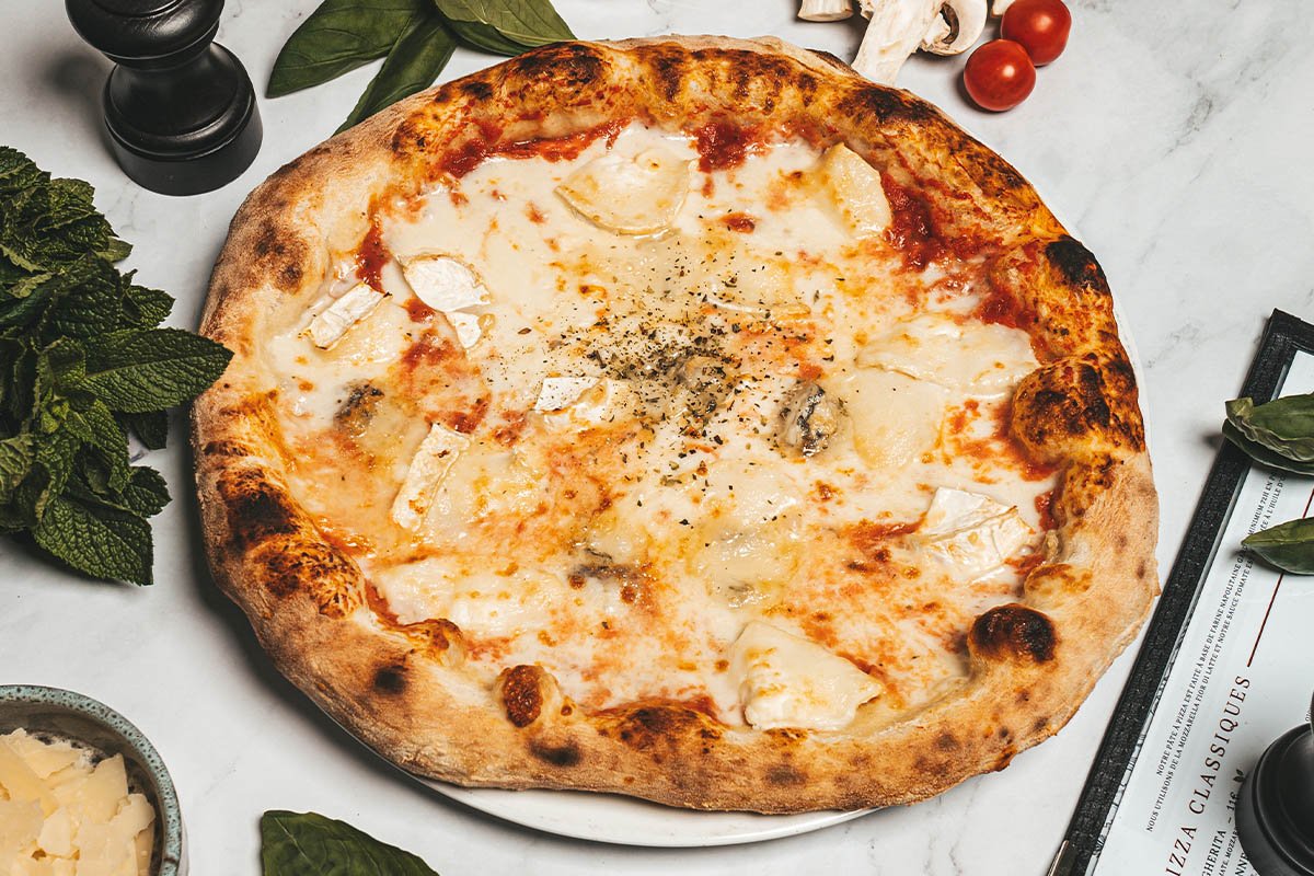 Une pizza blanche avec fromage, champignons et fines herbes sur une table en marbre blanche, entourée d'herbes, de tomates cerises, de sel et de poivre.