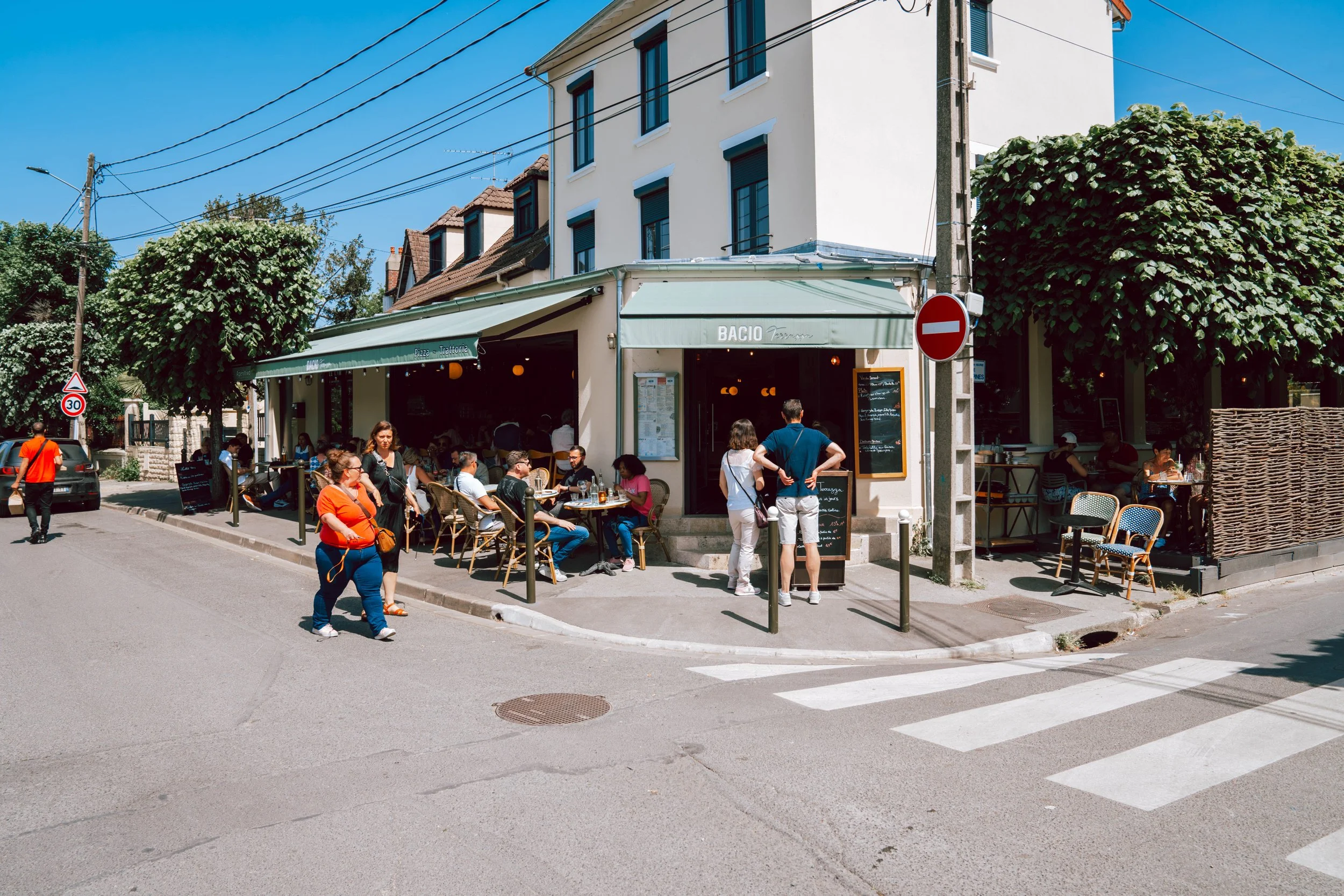 Un café en plein air avec des clients assis à des tables sous une enseigne portant le nom "BACIO". Des personnes attendent à l'entrée et d'autres se promènent dans la rue ensoleillée.