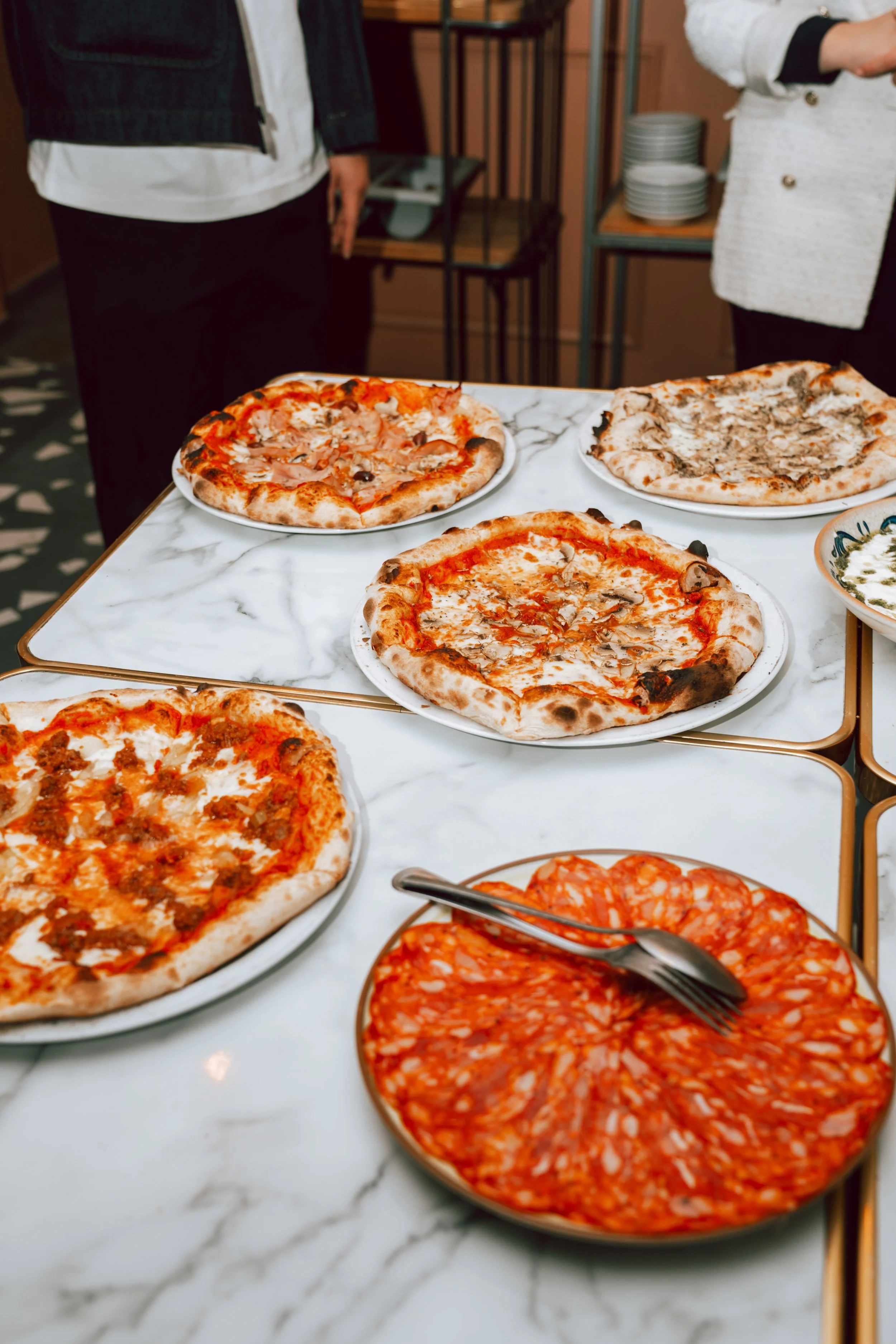 Plateaux de pizzas variées sur une table en marbre, avec des personnes en arrière-plan.