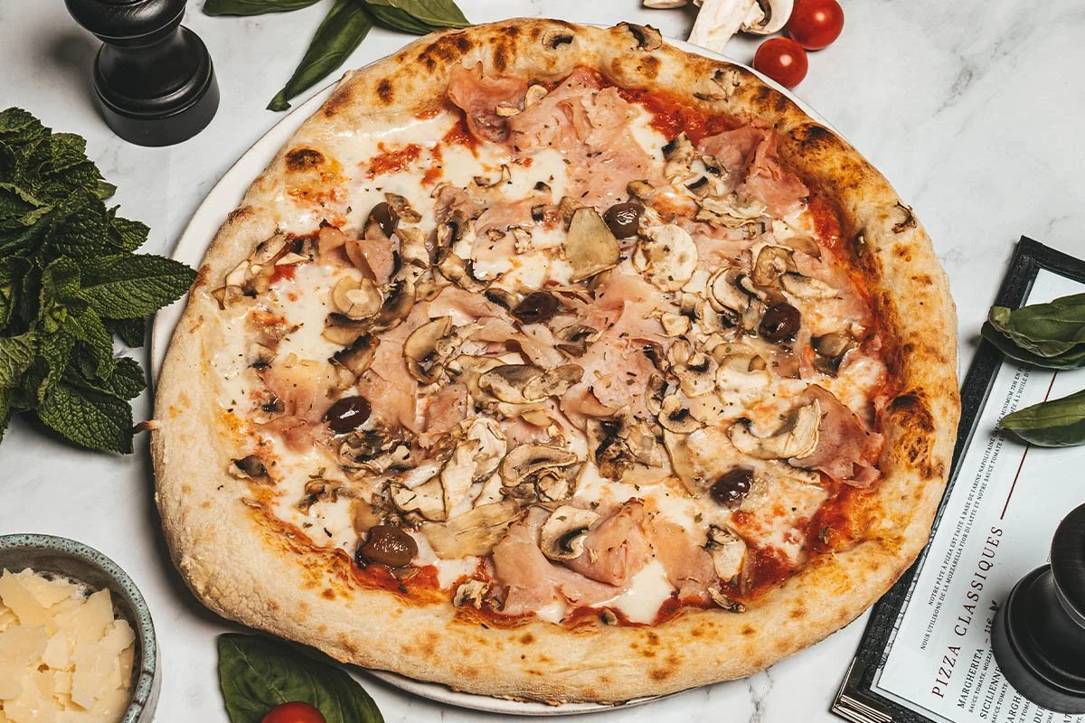Une pizza aux champignons, jambon et olives sur une assiette à côté de tomates cerises, de basilic, de menthe et de parmesan râpé.