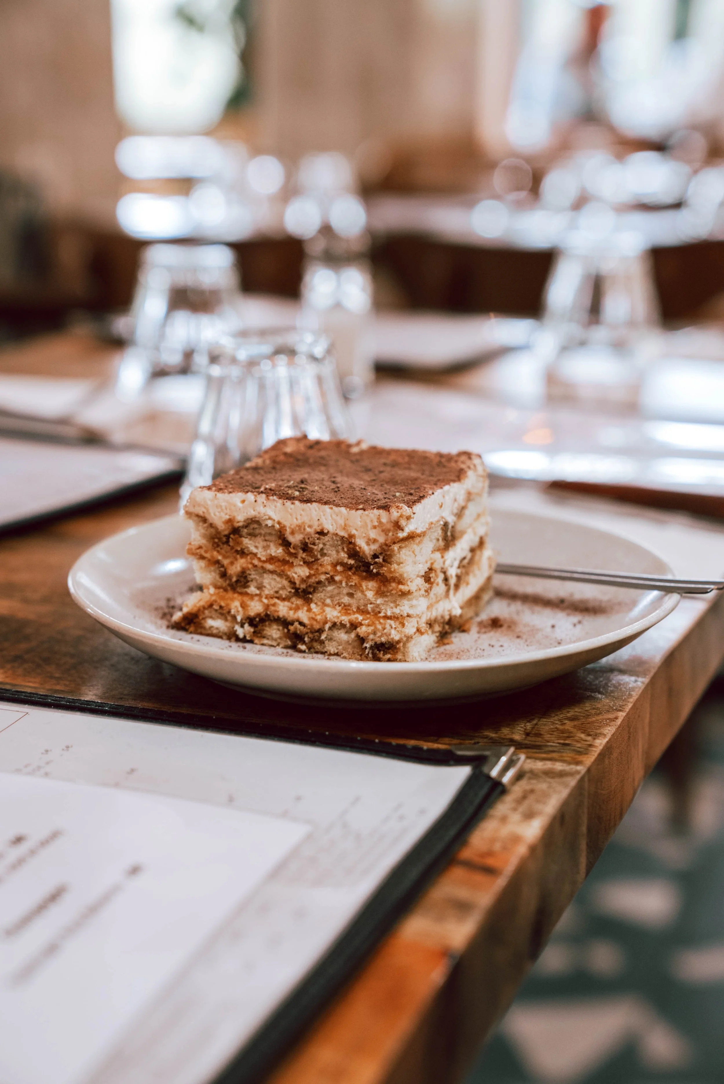 Une tranche de tiramisu posée sur une assiette blanche sur une table en bois, avec des verres à vin inversés et des éléments de décoration flous en arrière-plan.