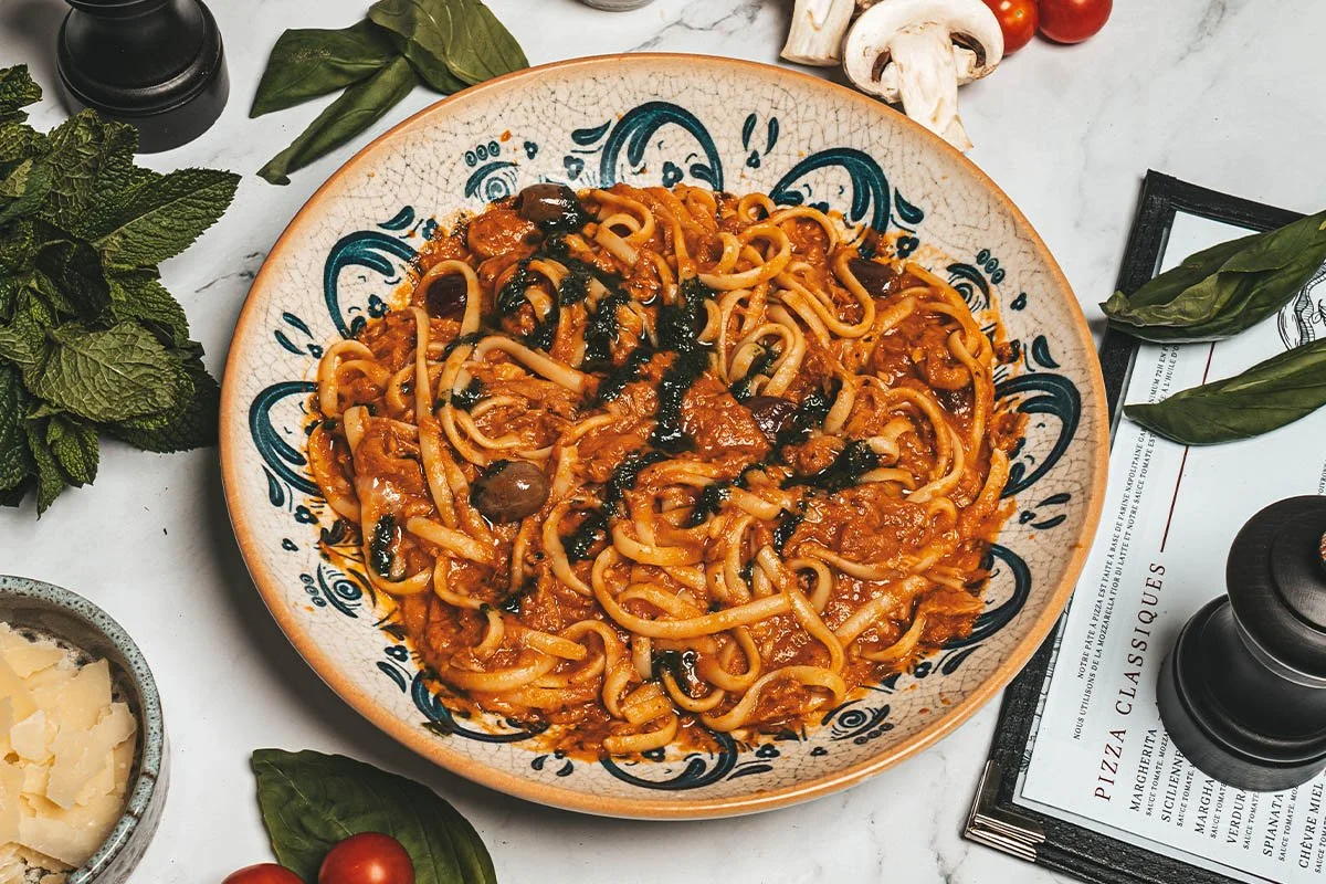 Plat de pâtes avec sauce tomate, olives noires, et légumes, dans un bol décoratif, entouré de basilic, champignons, tomates cerises, fromage, et condiments.