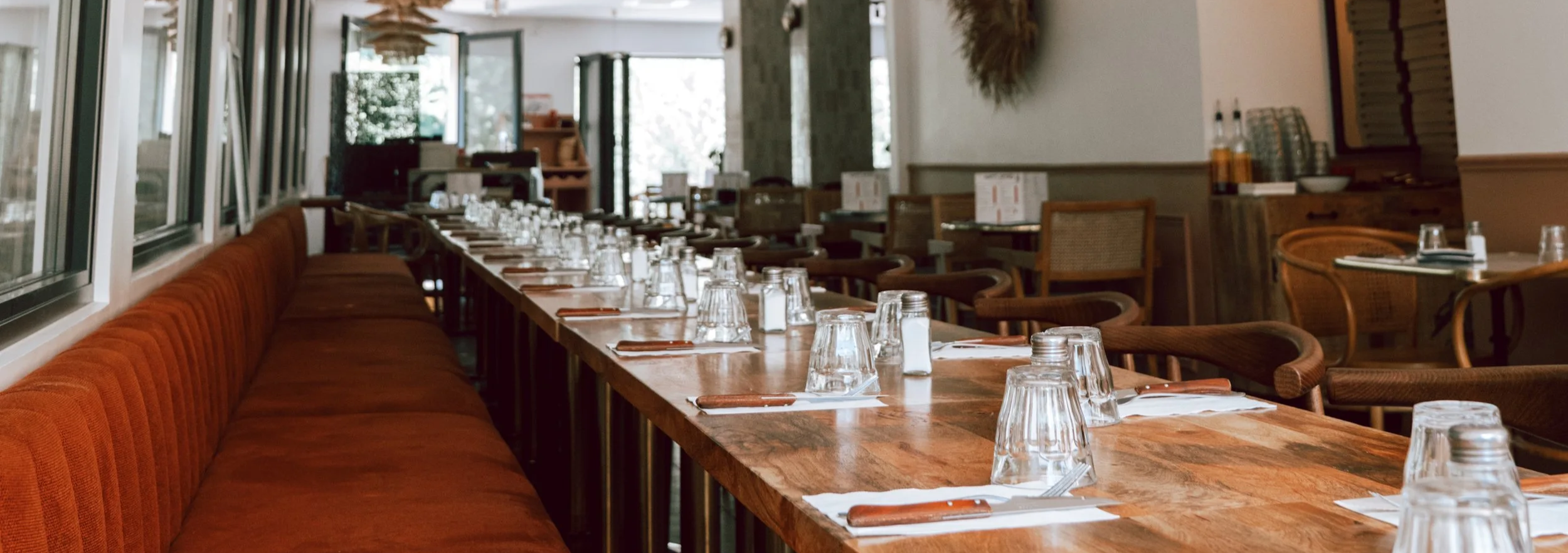 Salle à manger de restaurant avec table longue, verres à boire, assiettes, couverts, chaises en bois, décoration intérieure chaleureuse et grandes fenêtres.