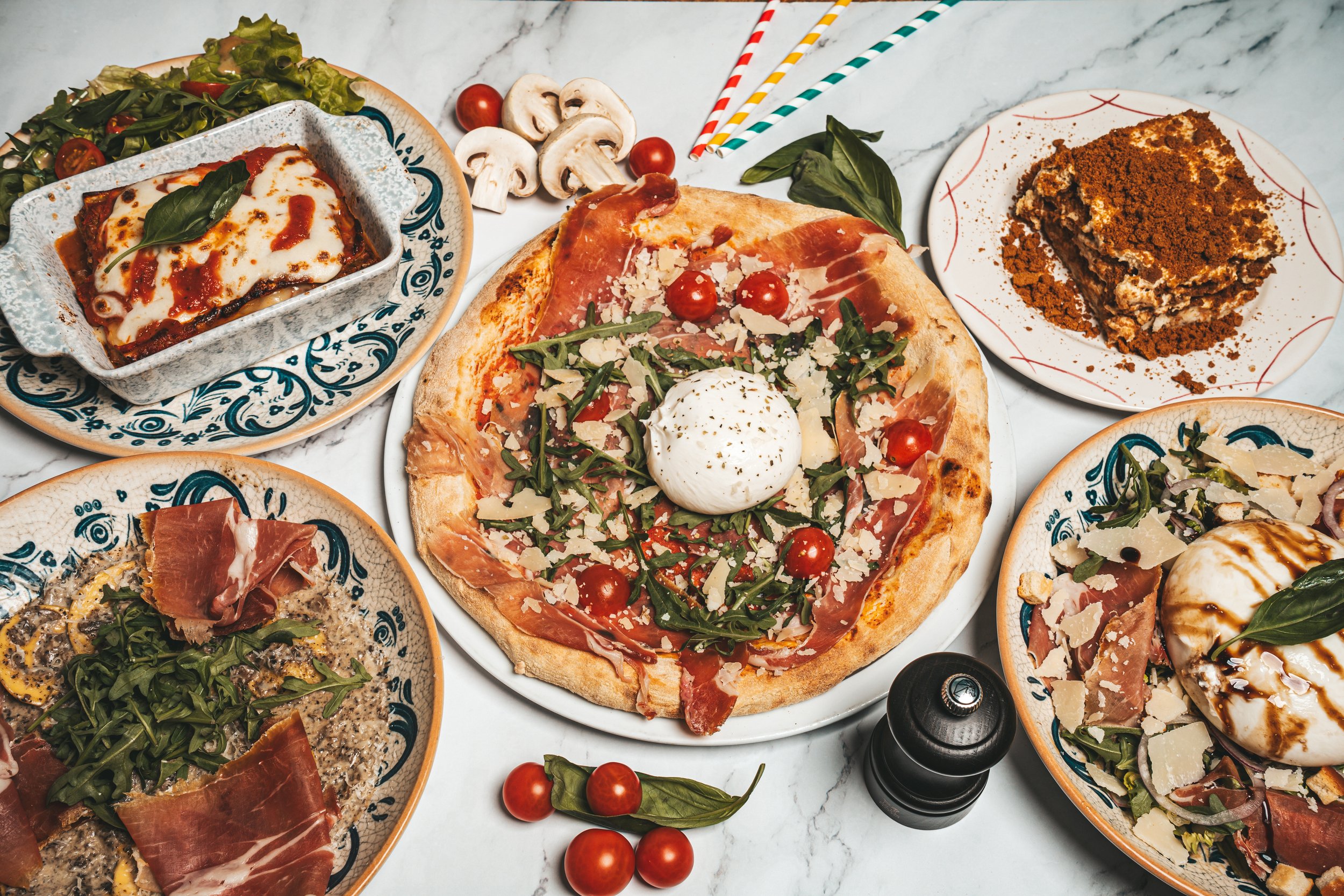 Une variété de plats italiens, notamment une pizza avec de la mozzarella, du jambon, des tomates cerises, et du basilic, accompagnée d'un plat de lasagnes, d'une part de tiramisu, d'une salade avec du jambon, de la roquette et du parmesan, le tout présenté sur une table en marbre.