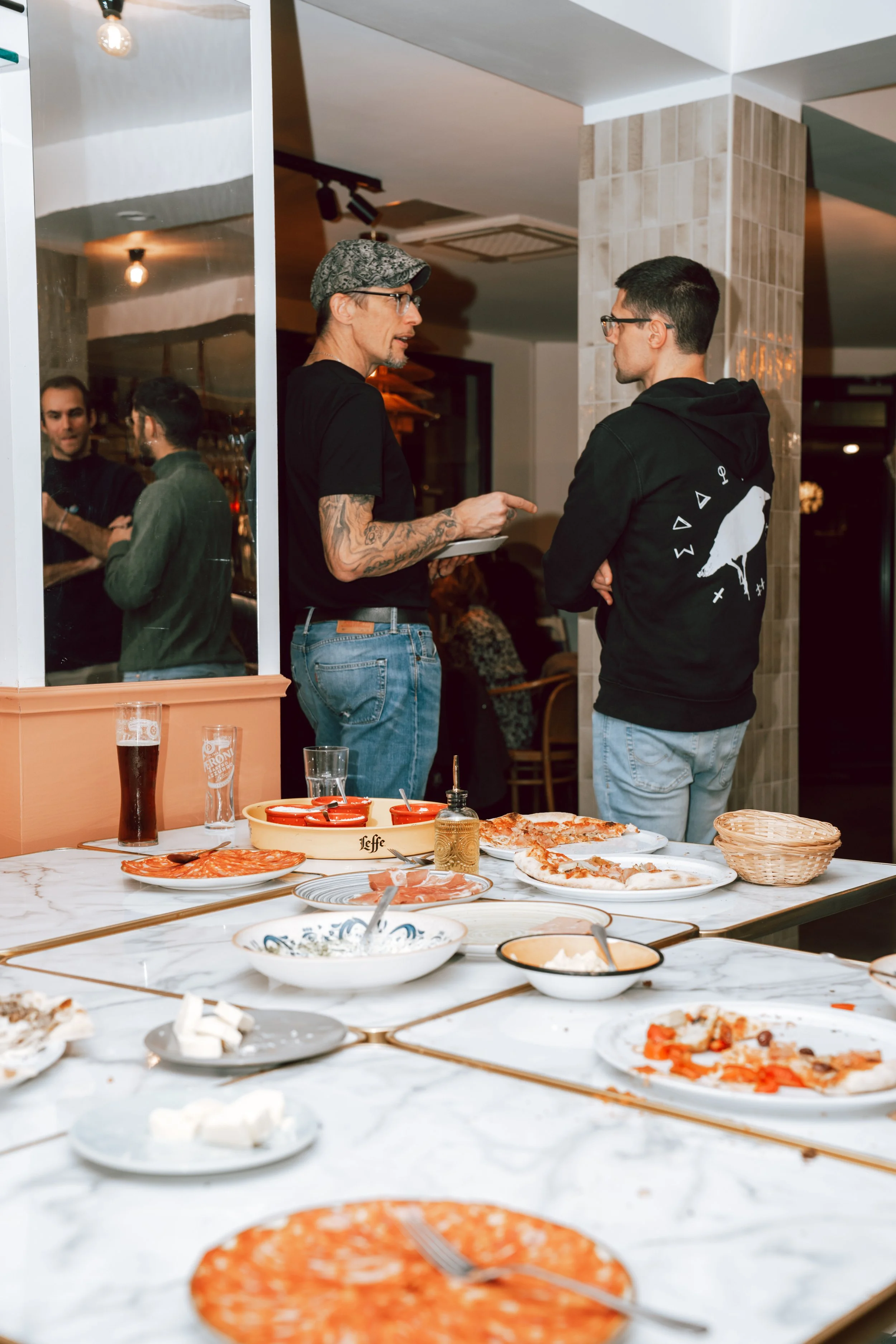 Deux hommes discutant dans un restaurant, avec des plats de pizza et de la nourriture sur la table en premier plan.