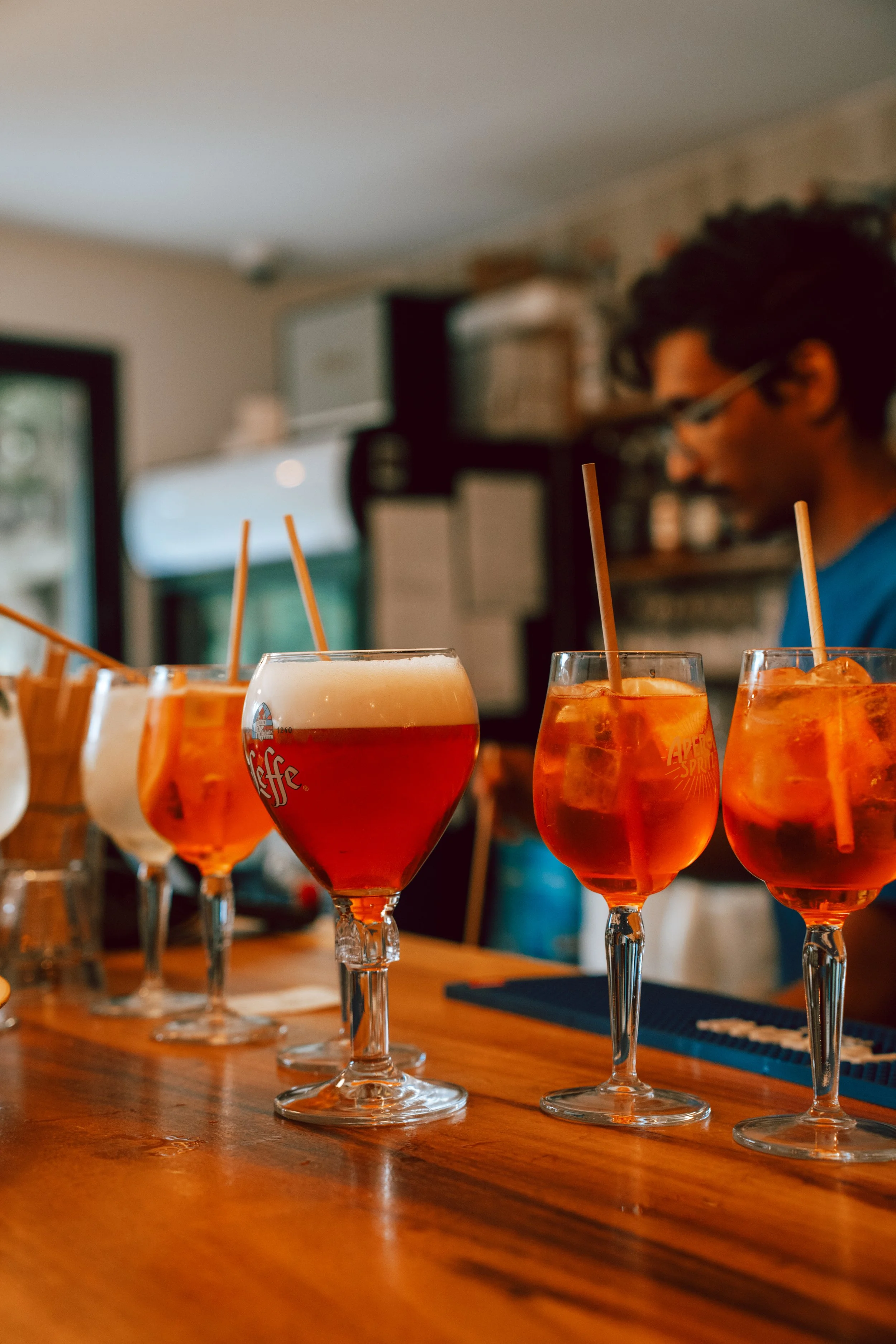 Verres à cocktails avec une boisson orange, straws en plastique, sur une table en bois dans un restaurant, personne en arrière-plan.