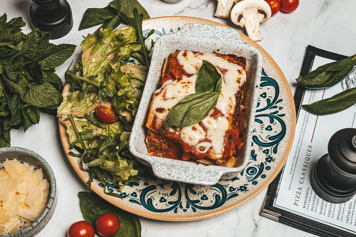 Une portion de lasagne à la mozzarella sur une assiette avec des feuilles de basilic, salade verte, tomates cerises, et Parmesan râpé sur la table.