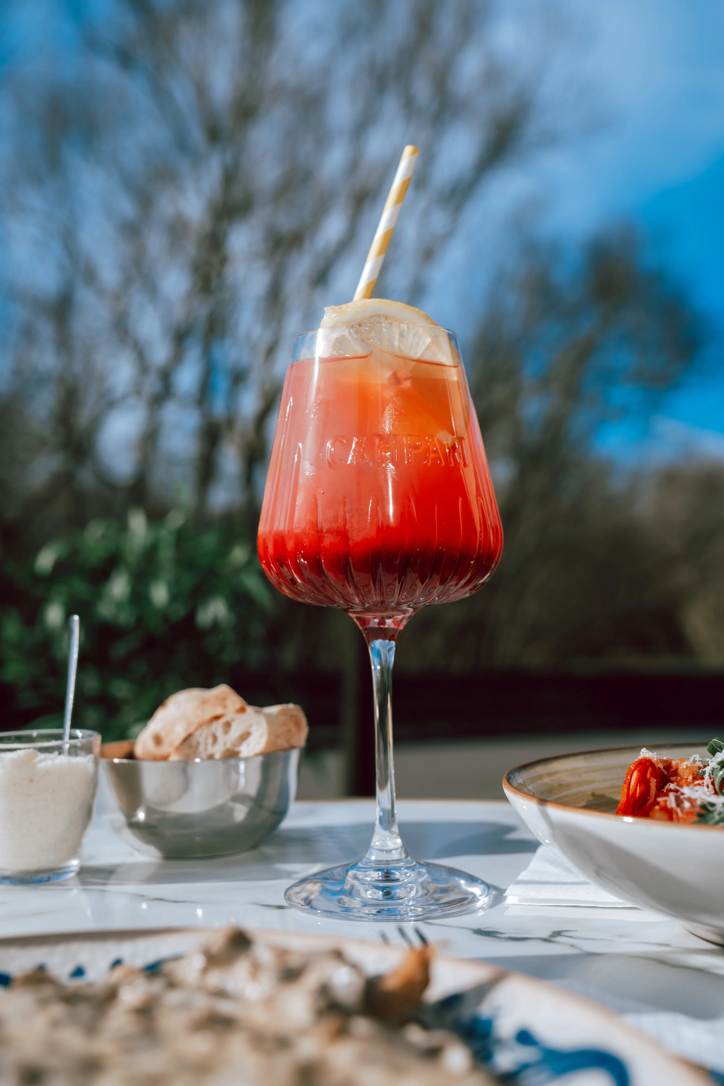 Verre à cocktail rouge avec tranche de citron, paille rayée beige et blanche, assiettes avec pain et plats de nourriture, fond de ciel bleu avec arbres dénués de feuilles.