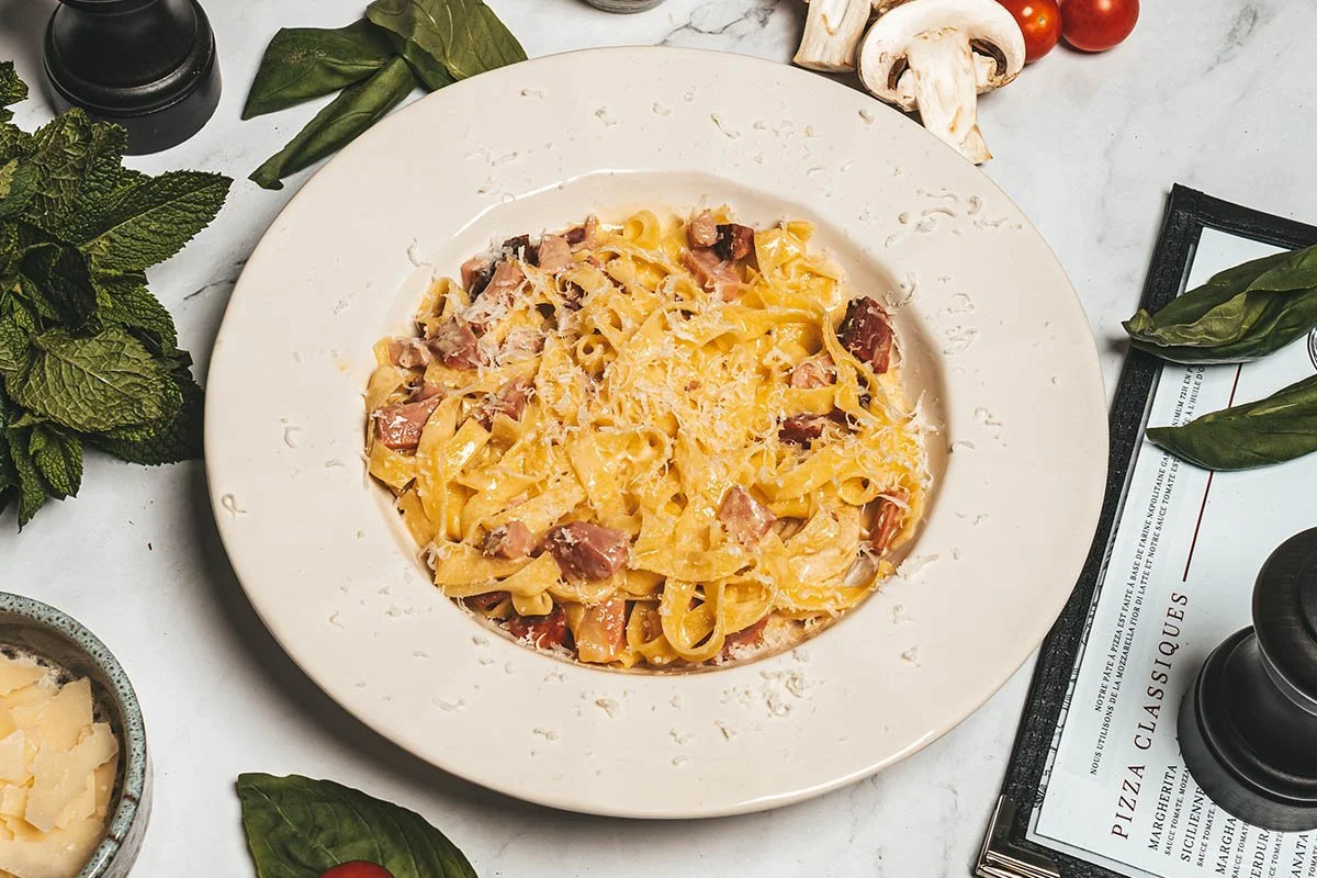 Plat de pâtes comme des tagliatelles avec des morceaux de jambon et du fromage râpé, présenté dans une assiette blanche.