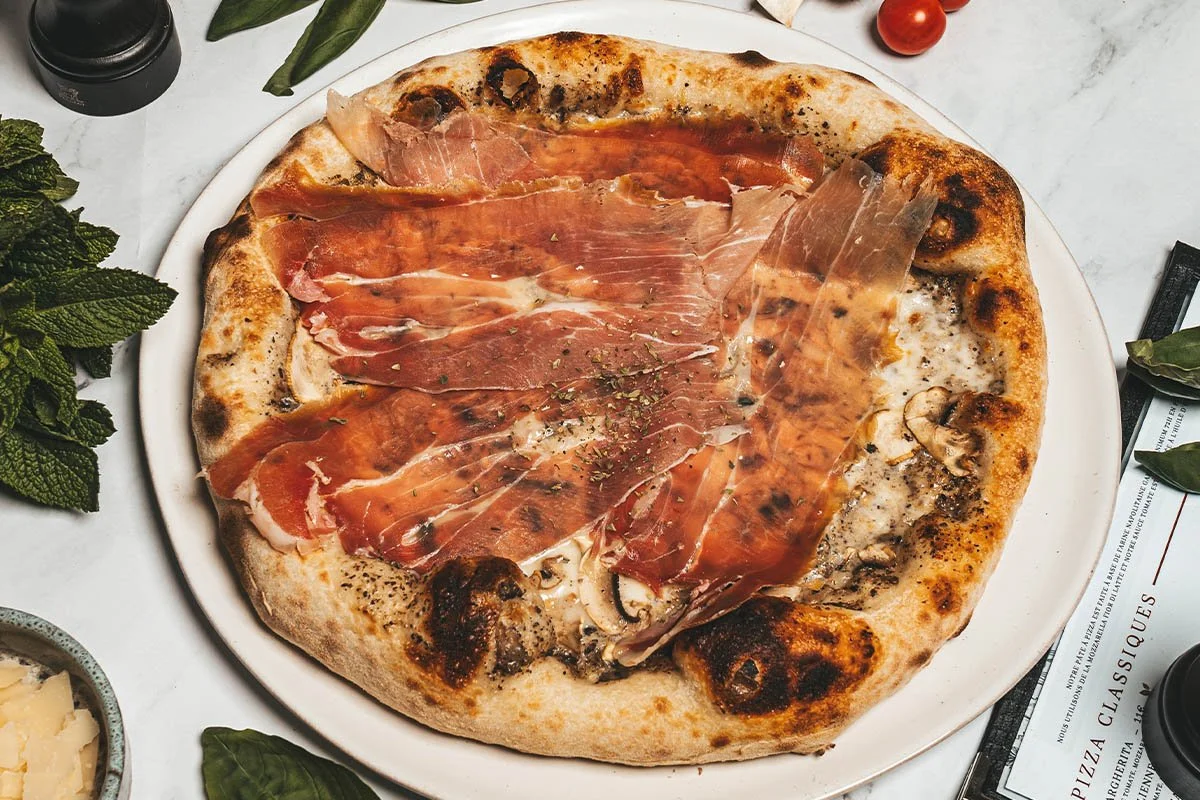 Une pizza garnie de jambon cru, champignons, fromage, et herbes, sur une assiette blanche, avec des feuilles de menthe, tomate, et des emballages à proximité.
