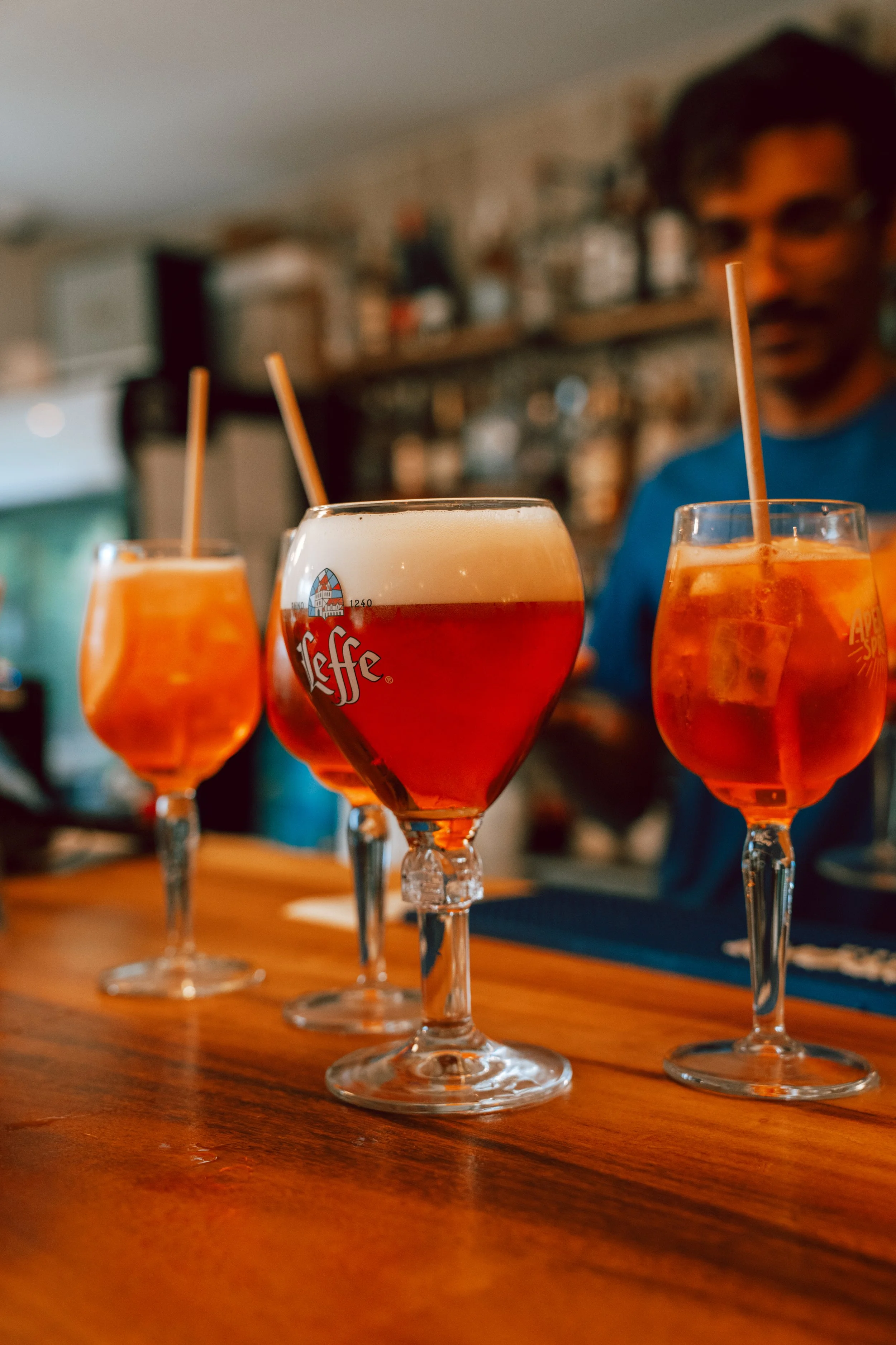 Trois verres de bière sur une table en bois dans un bar, avec une personne en arrière-plan. Le verre au centre affiche un logo "Leffe".