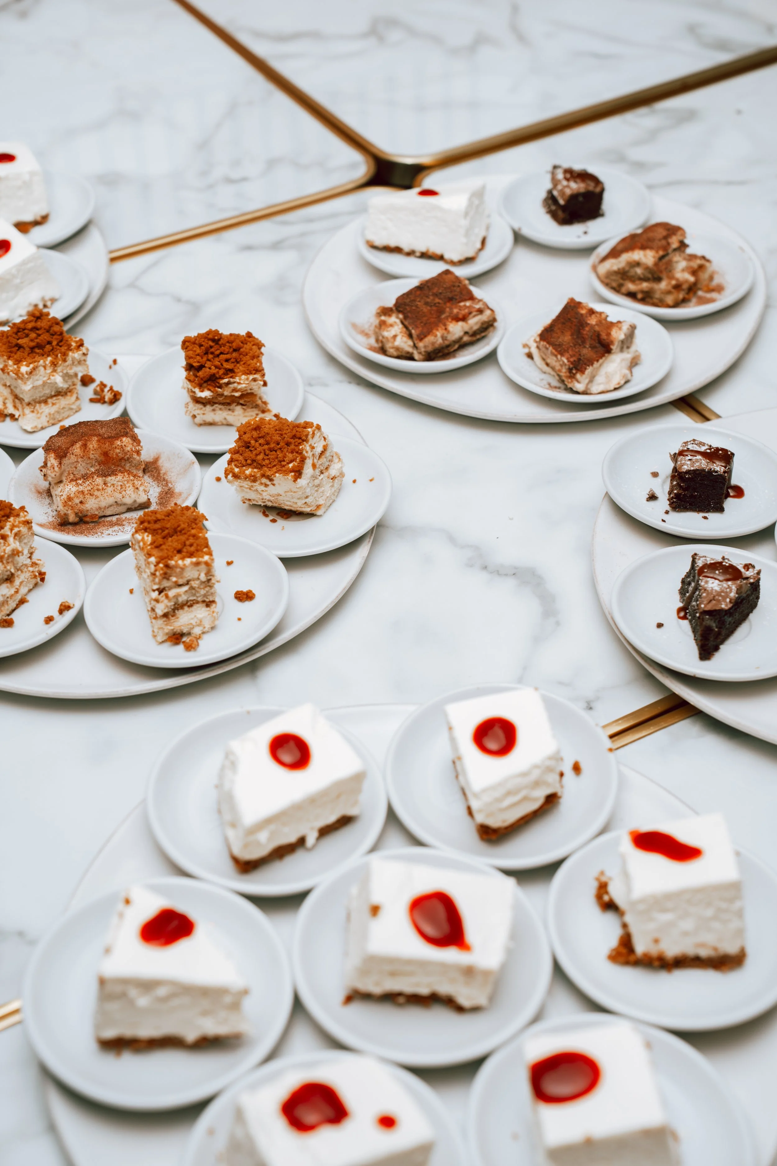Différents types de desserts sur des assiettes blanches posées sur une table en marbre, incluant des gâteaux avec des garnitures de crème, des morceaux de brownies, et des portions de gâteau au café avec une touche de coulis rouge.