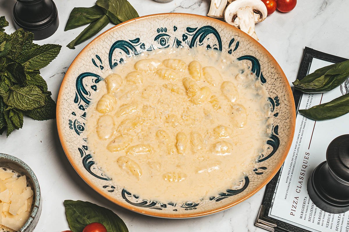 Baie de gnocchis dans une sauce crémeuse dans un bol décoratif, avec des herbes, champignons, tomates et autres ingrédients autour.