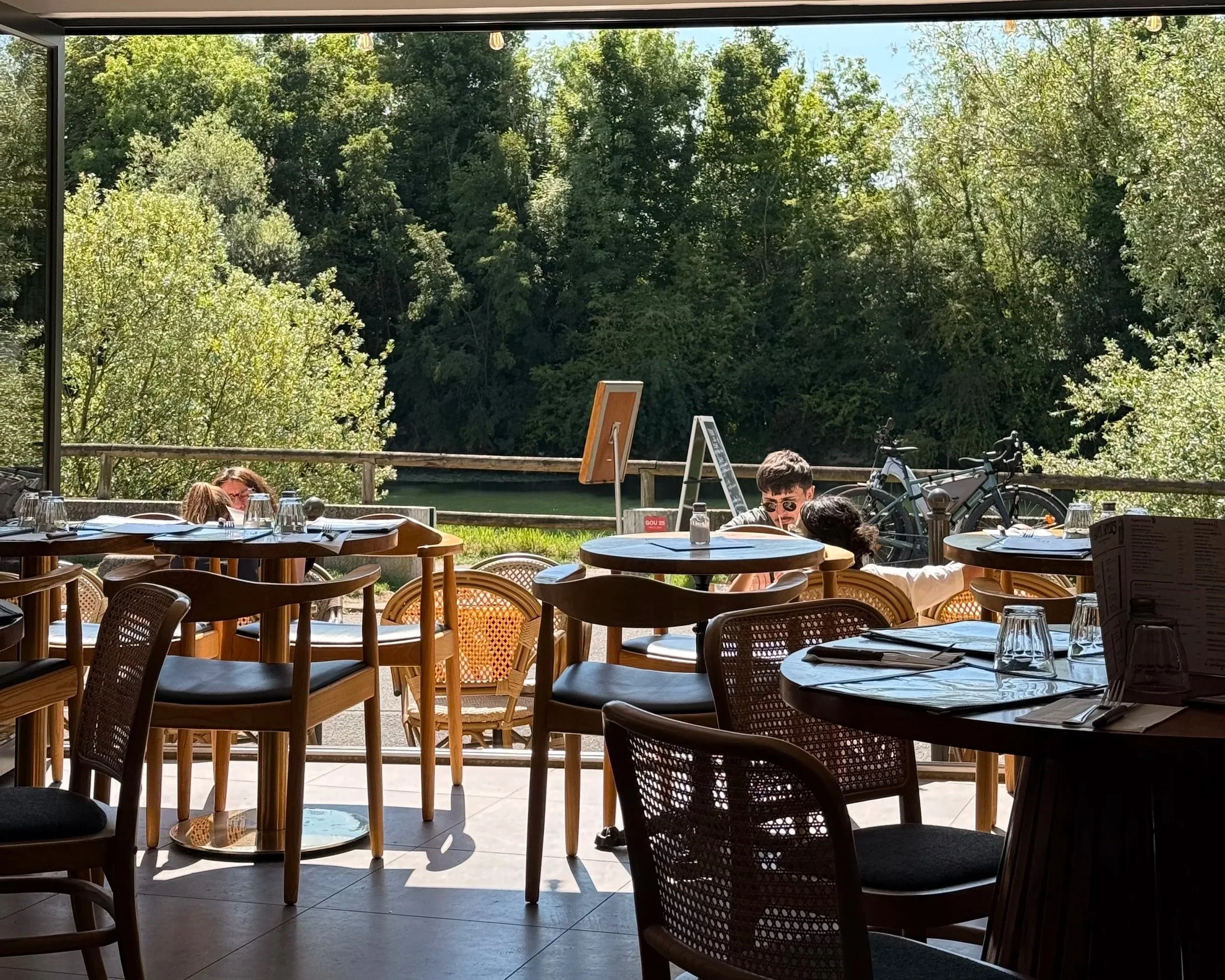 Terrasse de restaurant avec vue sur la nature, tables et chaises en bois, quelques personnes assises, bicyclette contre la rambarde, arbres verts en arrière-plan, restaurant en intérieur ouvert sur l'extérieur.
