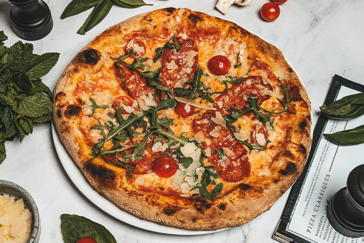 Une pizza avec fromage, tomates cerises, pepperoni, roquette et copeaux de parmesan, sur une assiette blanche sur une table en marbre.
