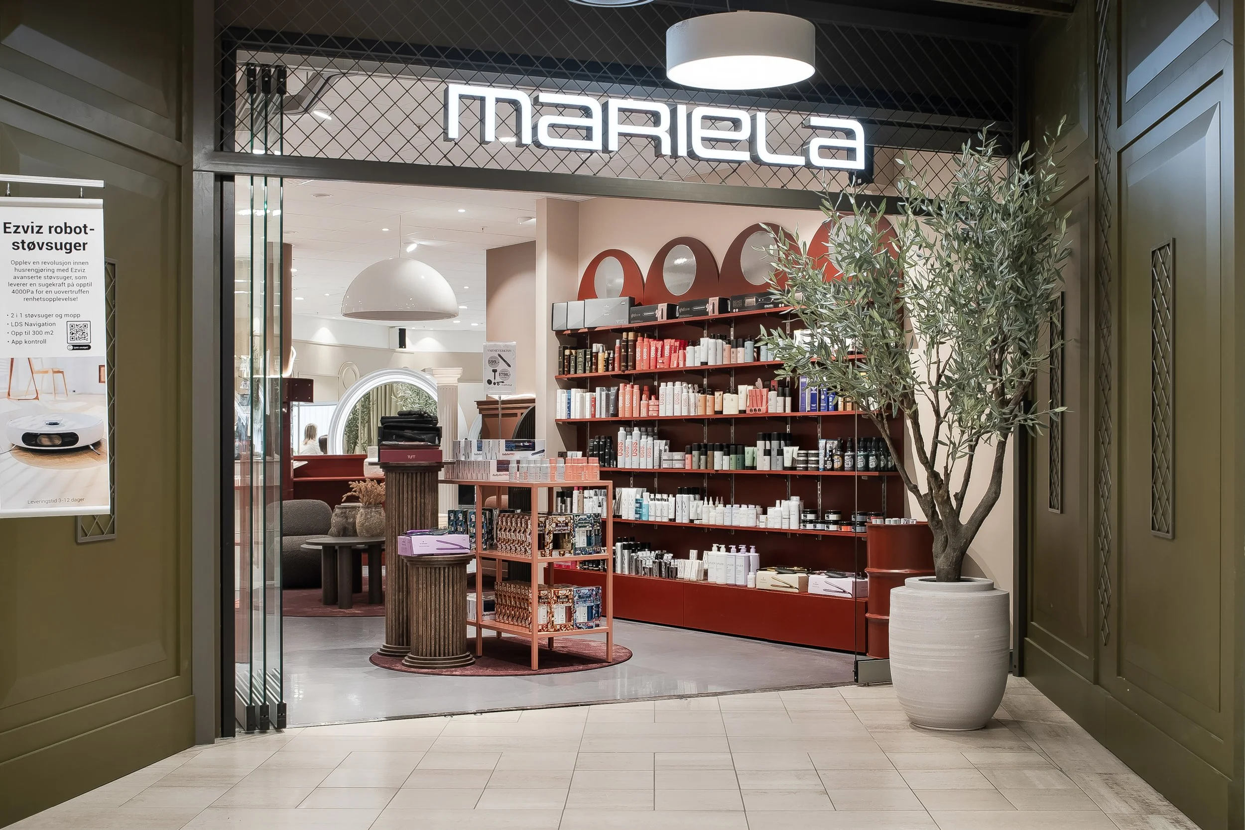 Innvendig butikk med mange produkter på hyller, en stor plante i en hvit potte, og et forretningsskilt som sier 'mariela'.