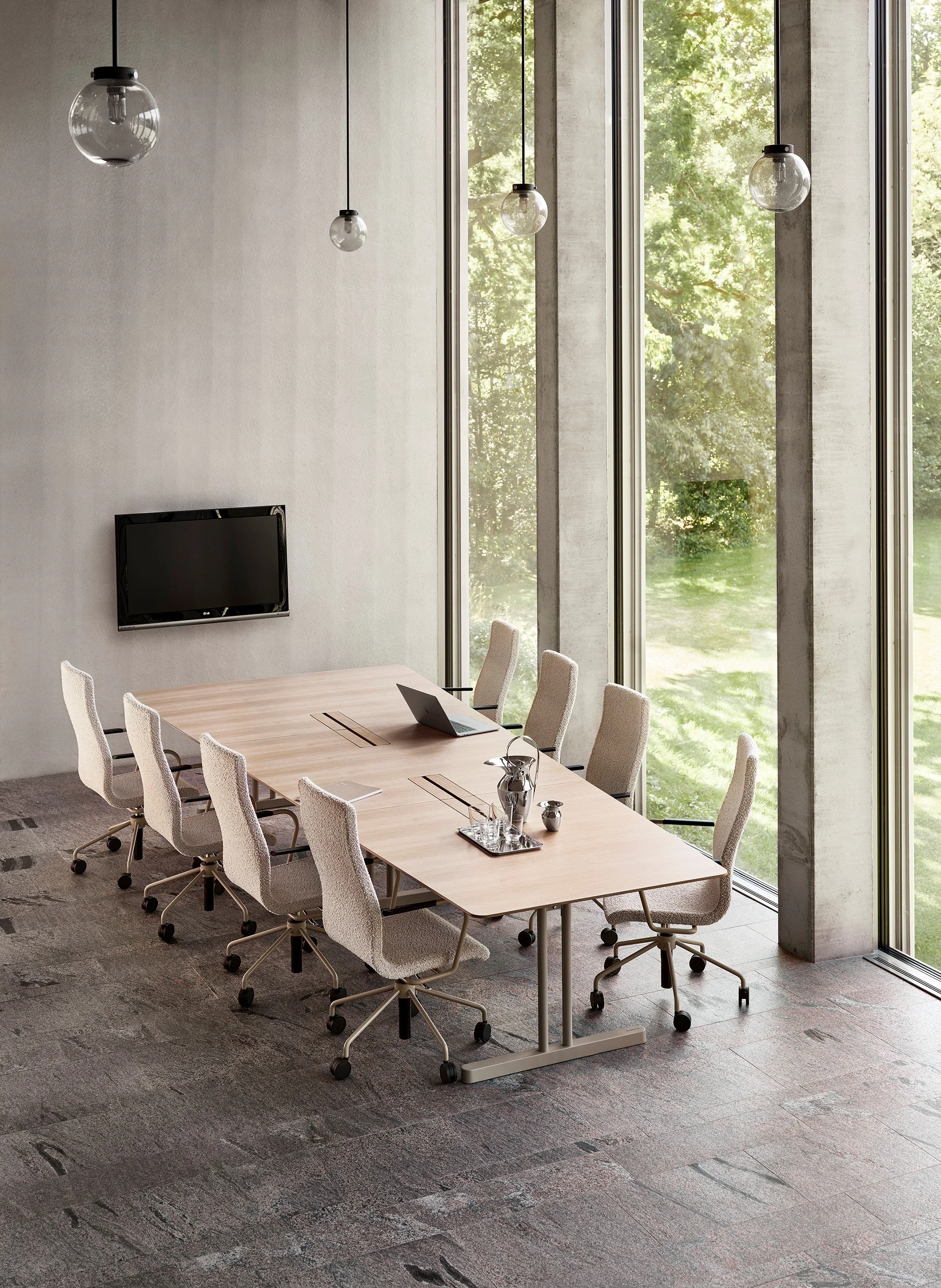 Konferanserom med stort bord, ni stoler, store vinduer med utsikt over grønt naturområde, tre glasslamper henger i taket, flatskjerm på veggen, laptop og vaser på bordet.