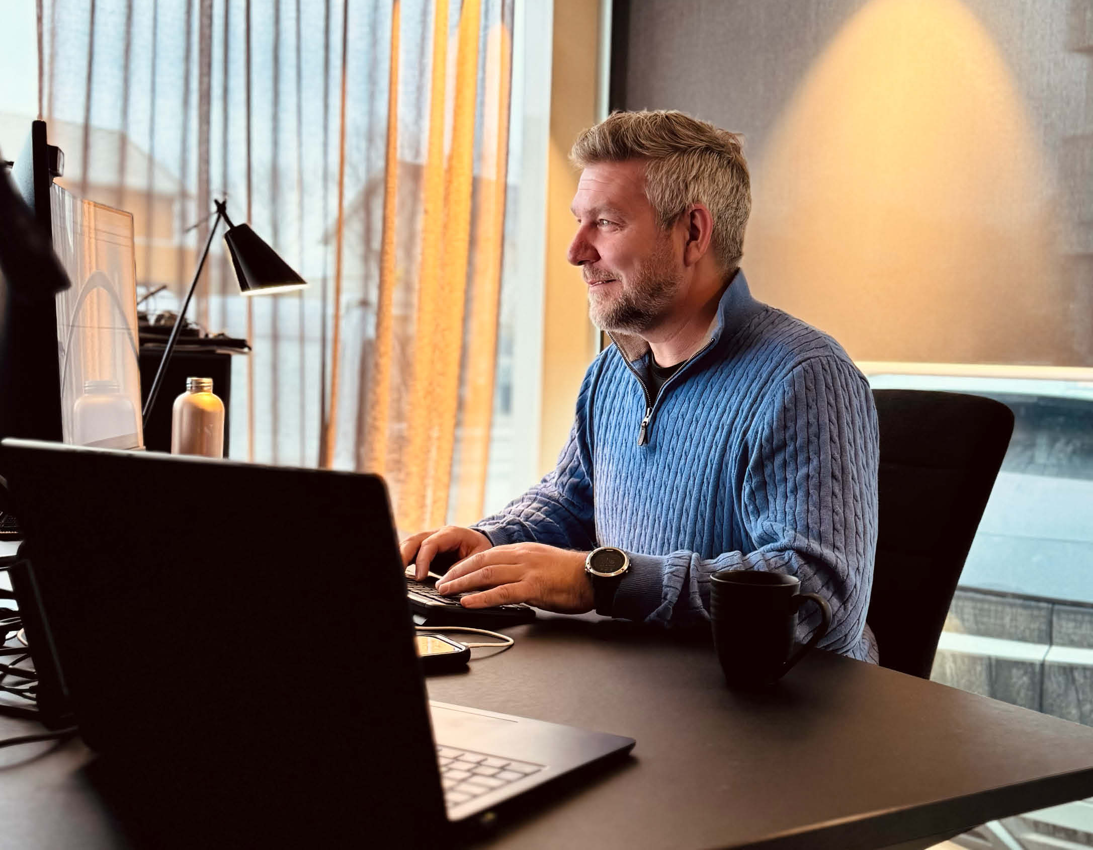 En mann med grått hår og skjegg som jobber foran en datamaskin på et kontor, med lys treverk og store vinduer.