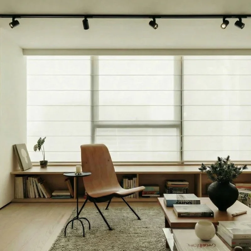 Et minimalistisk stue med stor vindusdekorasjon med blinds, en lenestol, et lite rundt bord, og en bokhylle under vinduet fylt med bøker. Det er en ryddig og lys atmosfære.