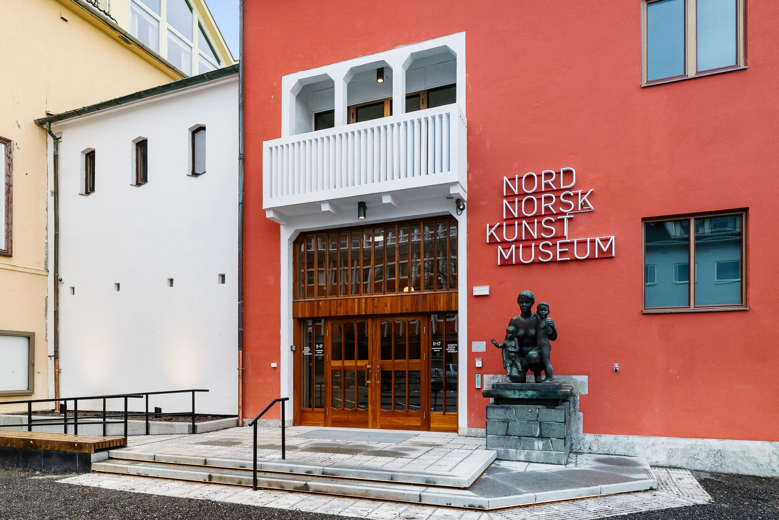 Bild av Nord Norsk Kunstmuseum med en rød fasade, en inngang av tre, en syre med en skulptur av to barn foran.