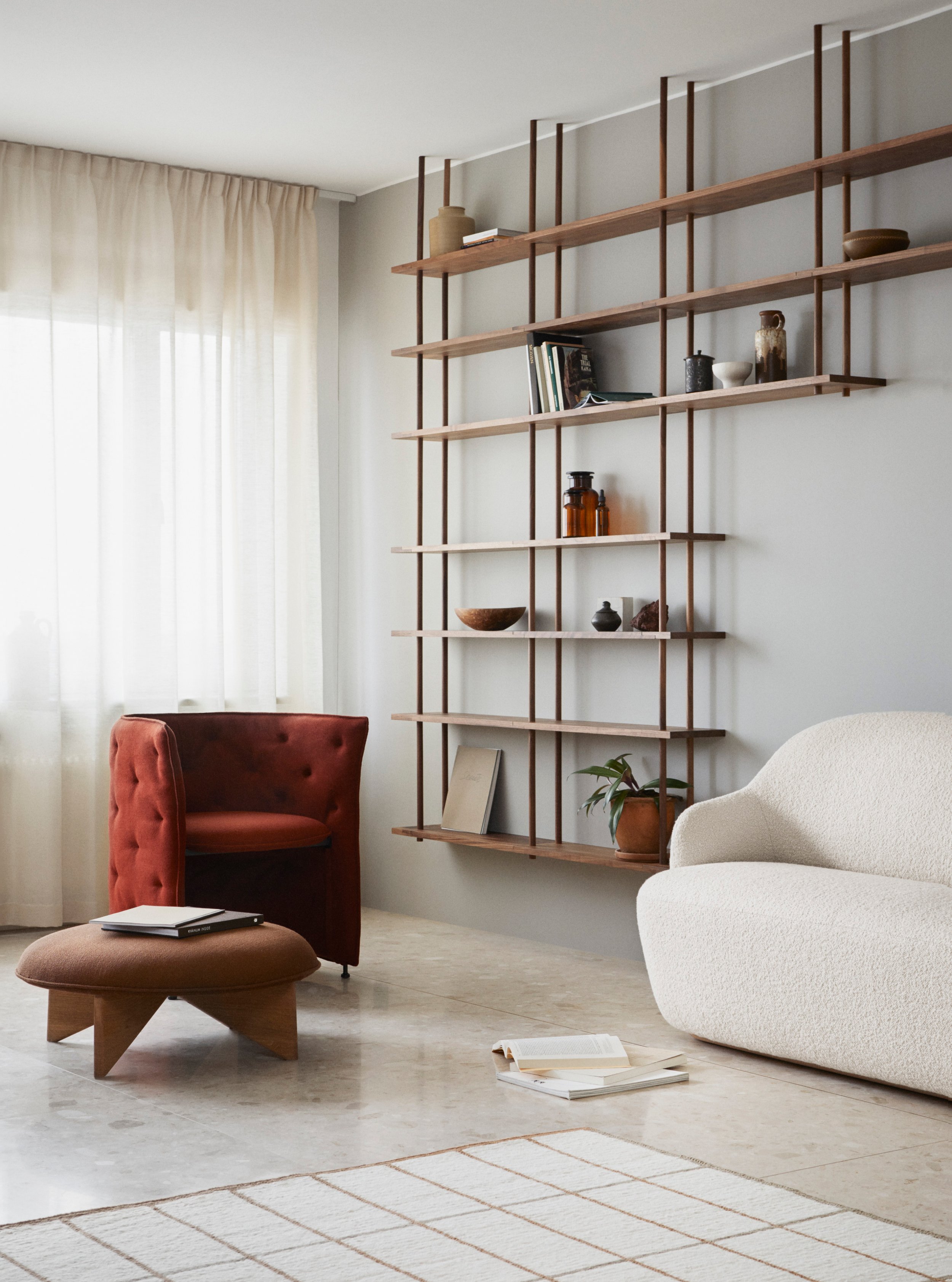 Et lyst og moderne stueinnredning med en romslig sofa, en rustfarget armstol, en responsiv pute, en åpen roterende bokhylle på veggen med dekorative gjenstander, og et mønstrete teppe på gulvet. Naturlig lys slipper inn gjennom vindu med hvite gardiner.