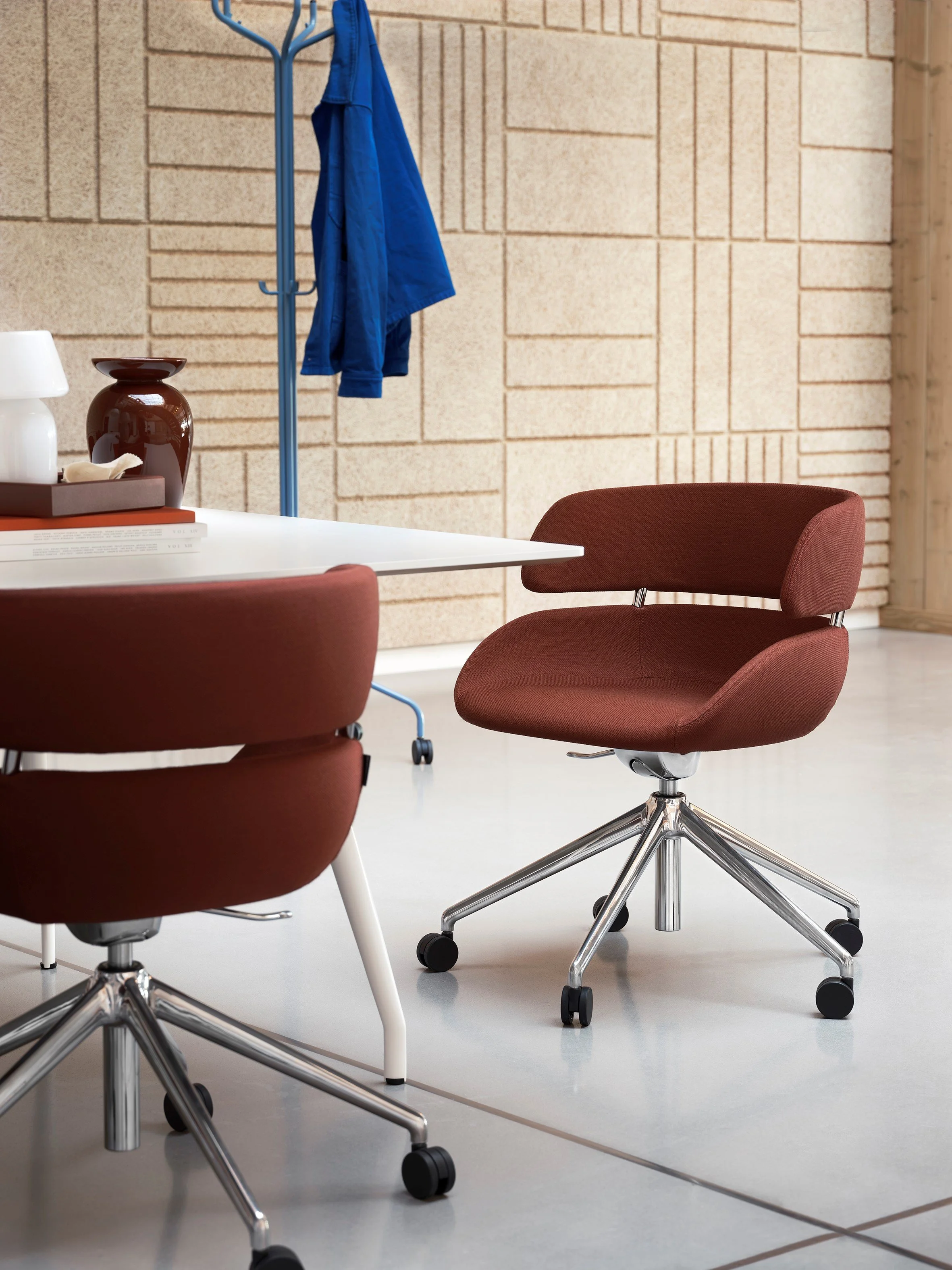 Kontorrom med røde kontorstoler, hvitt bord, og en kleshenger med en blå jakke, på en vegg med dekorative murstein.