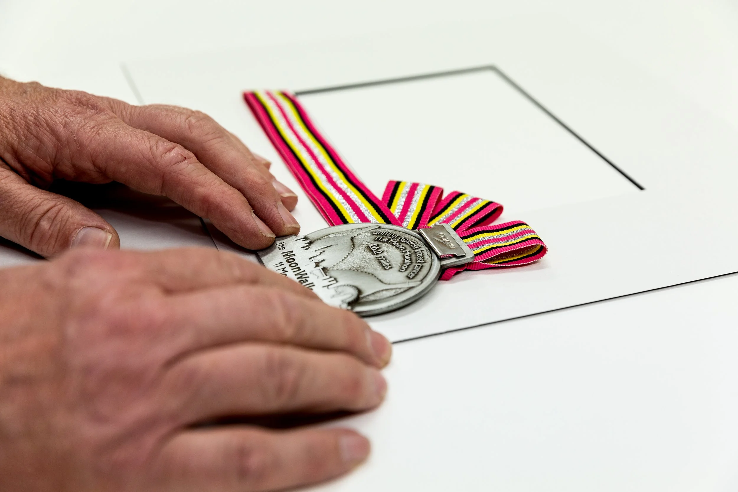 Person's hand placing a medal with a pink, yellow, and black striped ribbon onto a white surface.