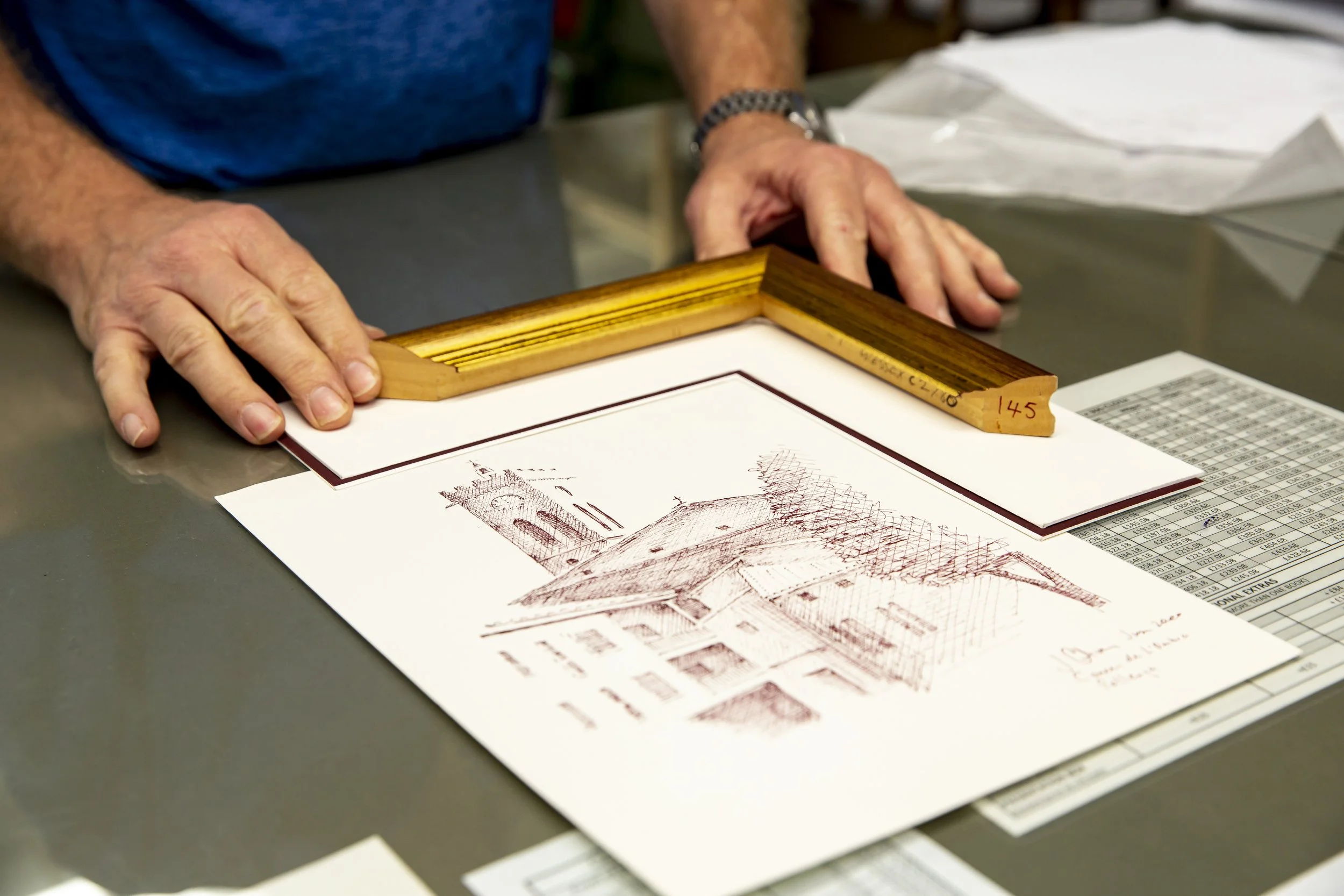 Person handling framed artwork or document on table with printing materials nearby.