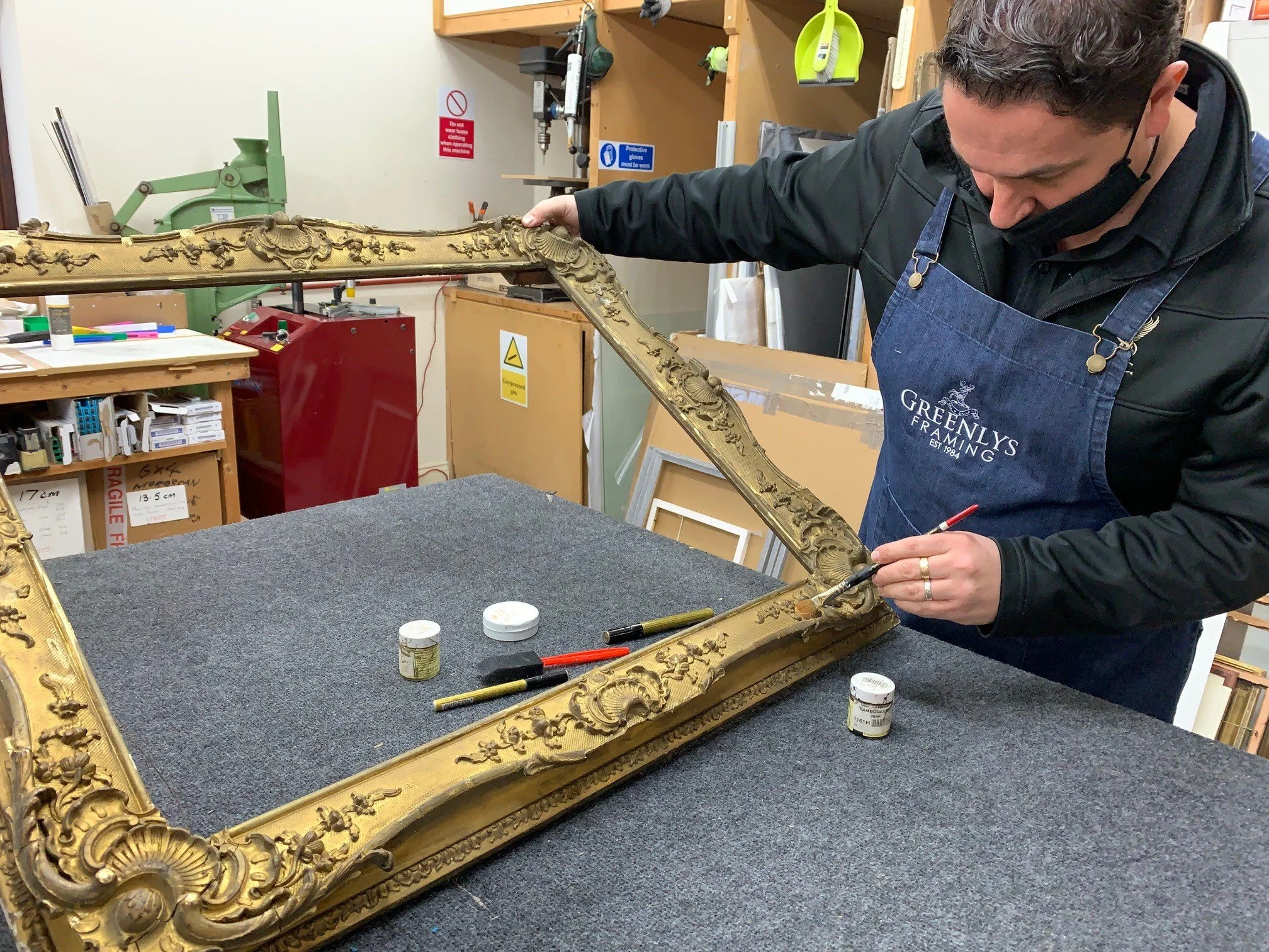 A person wearing a face mask and apron working on an ornate, gold-colored, carved picture frame in a workshop. The workshop is equipped with tools, paint, and safety signs.