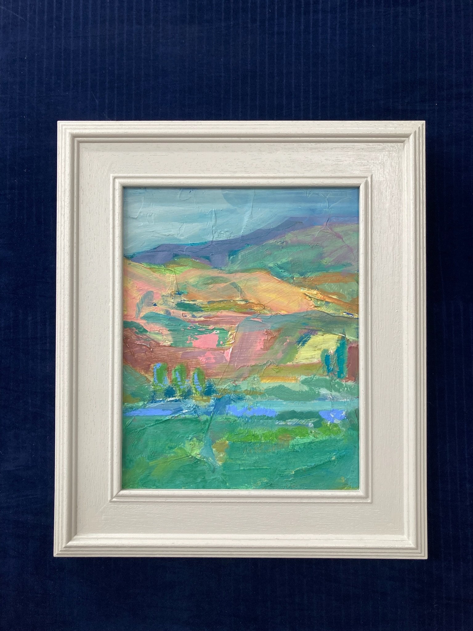 Abstract landscape painting with mountains, sky, and lush green foreground, framed in white, displayed on a dark blue wall.