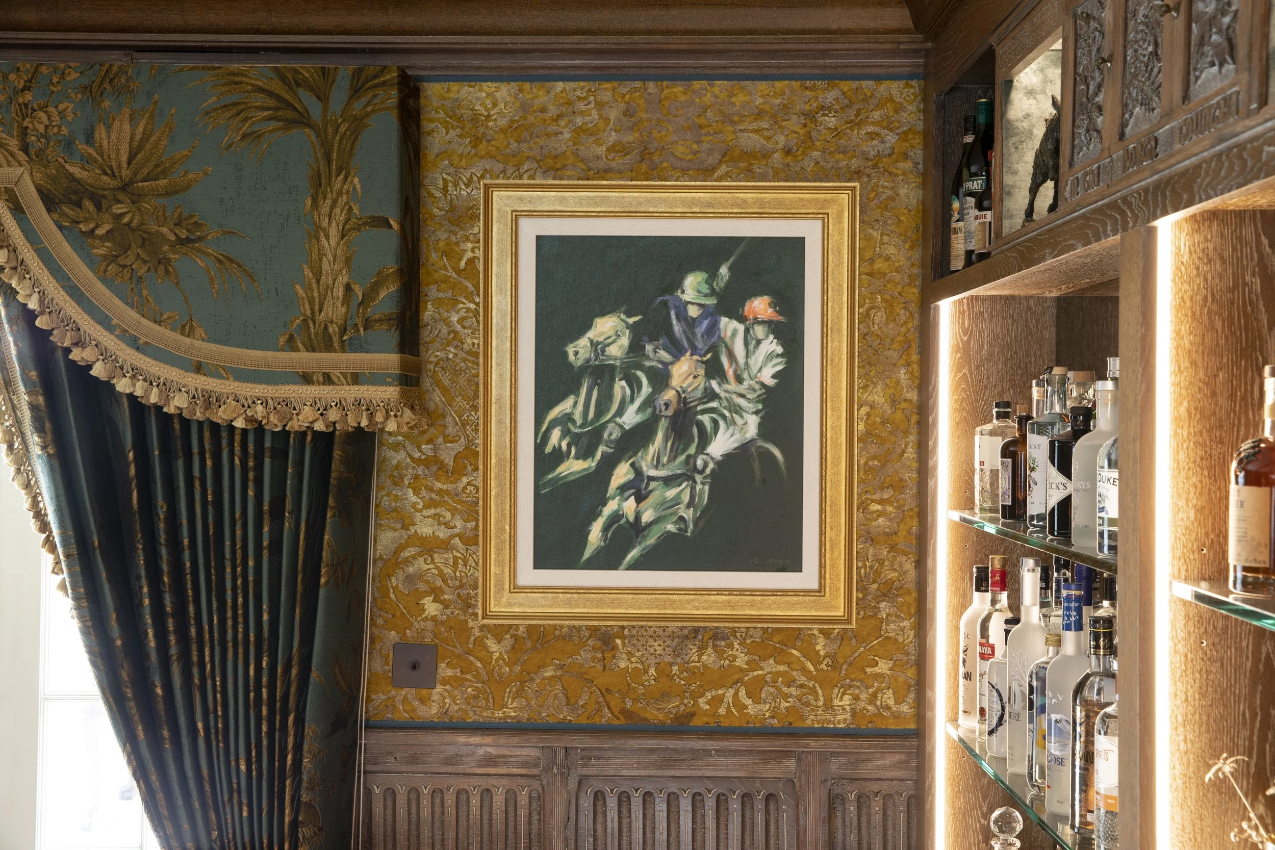 Interior corner of a room with ornate wallpaper, a framed painting of jockeys on horses, a dark wood shelf with bottles, a decorative curtain, and a window showing natural daylight.