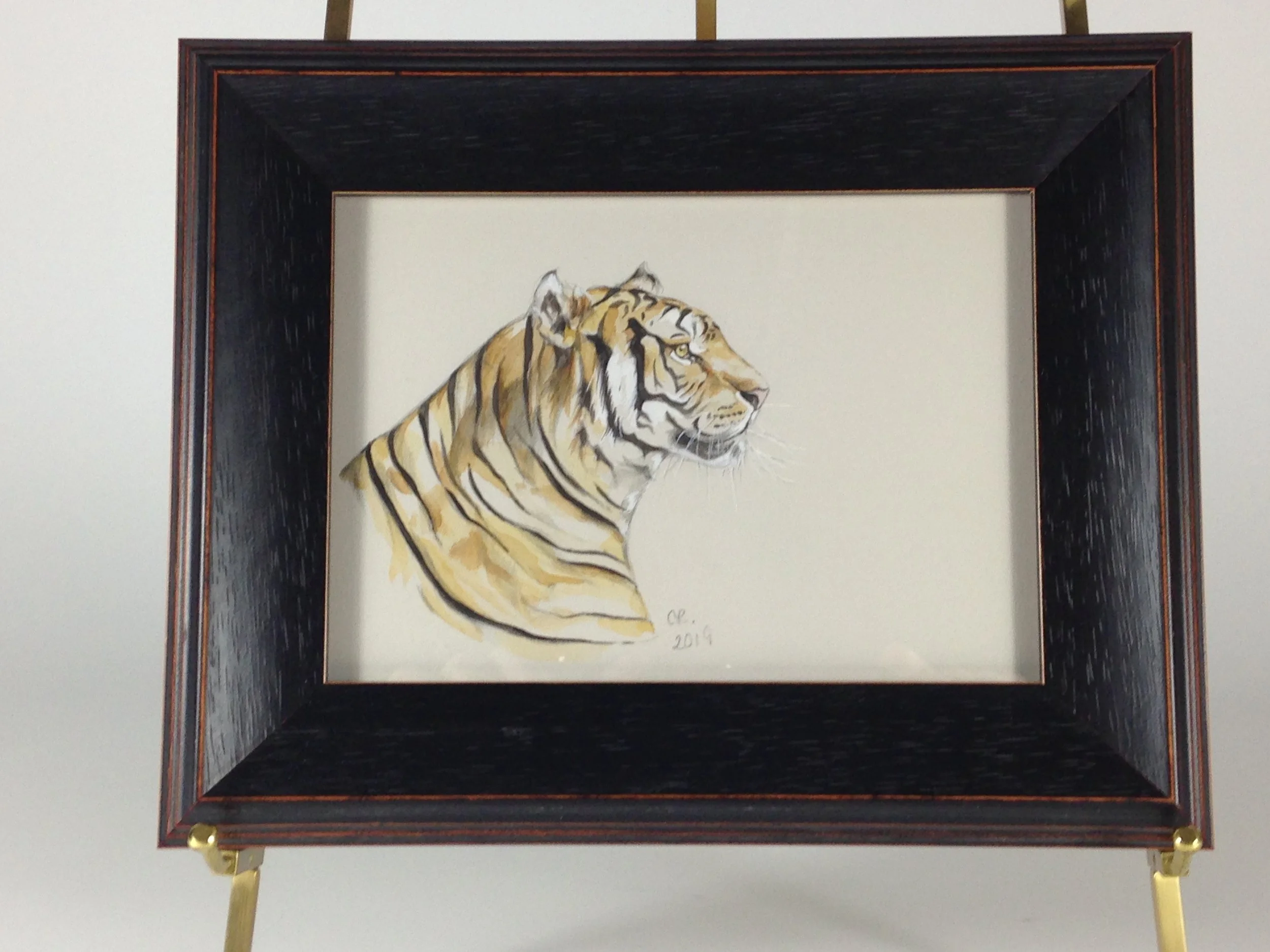 A framed watercolor painting of a tiger's head and upper body, signed by the artist, dated 2019, and on display with a stand and gold-colored fasteners.