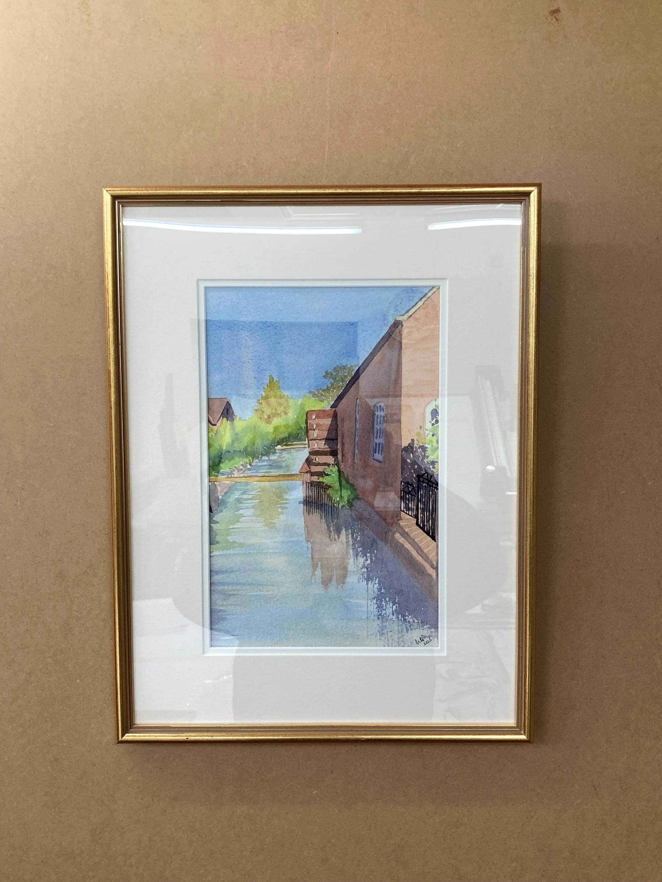 Framed watercolor painting of a river scene with buildings and trees on a wall.