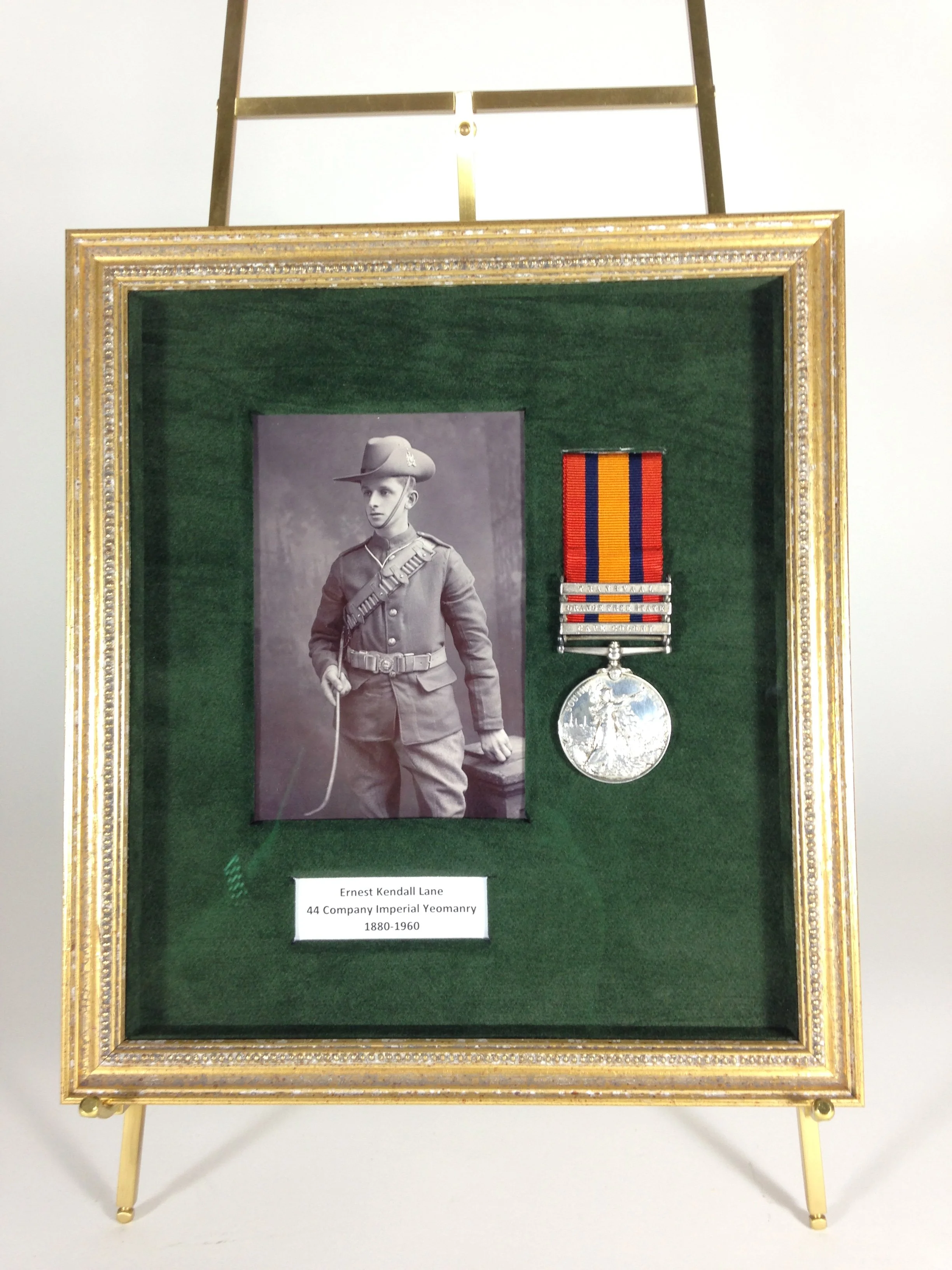 Display case with a black-and-white photograph of a soldier in uniform and a medal with a ribbon in red, blue, and orange colors. There is a small label beneath the display that reads: 'Ernest Kendall Lane, 44 Company Imperial Yeomanry, 1880-1960.'