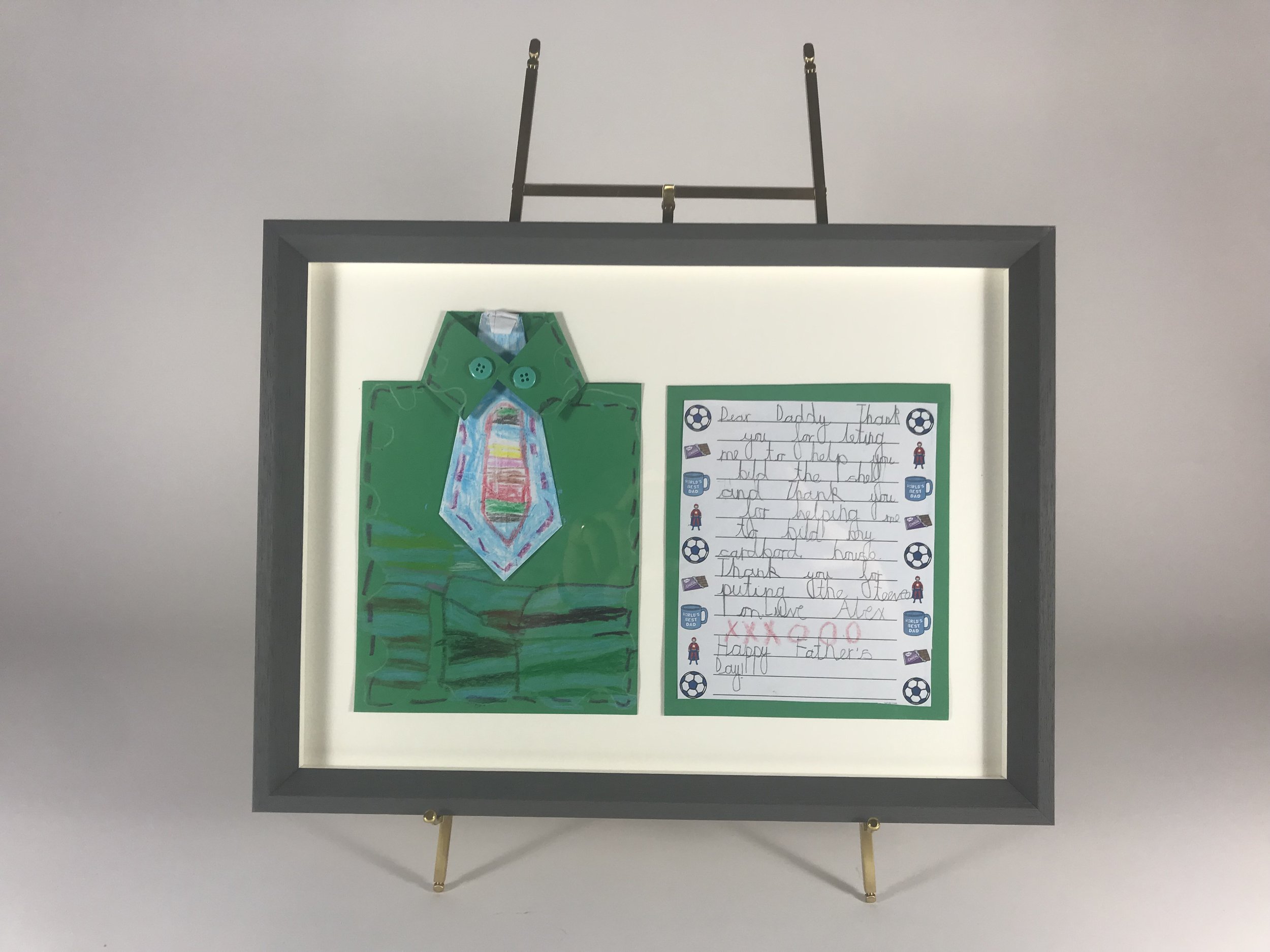 A framed display featuring a colorful, hand-drawn shirt and a handwritten note on a white paper, decorated with soccer ball stickers.