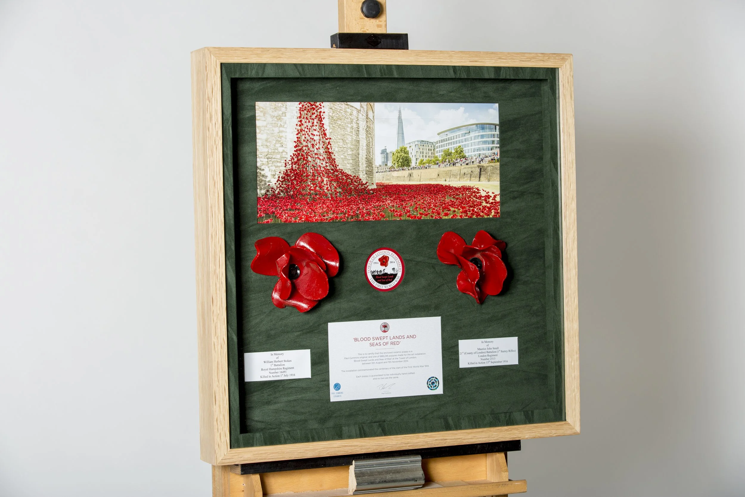 A framed photograph of a memorial display showing a field of red poppies, with some poppies attached to the frame below. The background of the photograph features a stone tower, modern buildings, and a cityscape. The display includes plaques and symb