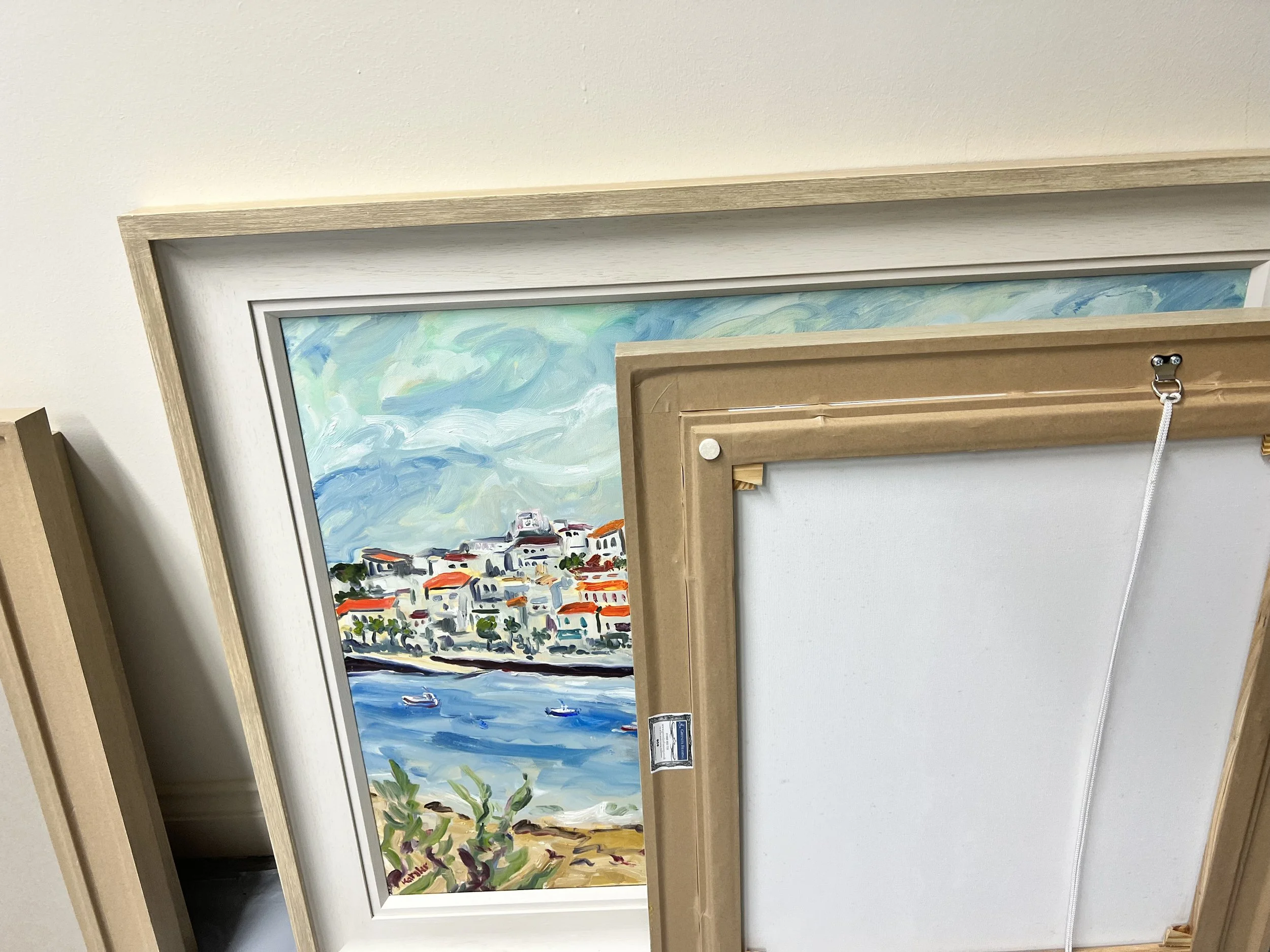 Three framed paintings hung vertically on a white wall in an interior space, each depicting colorful seaside scenes.