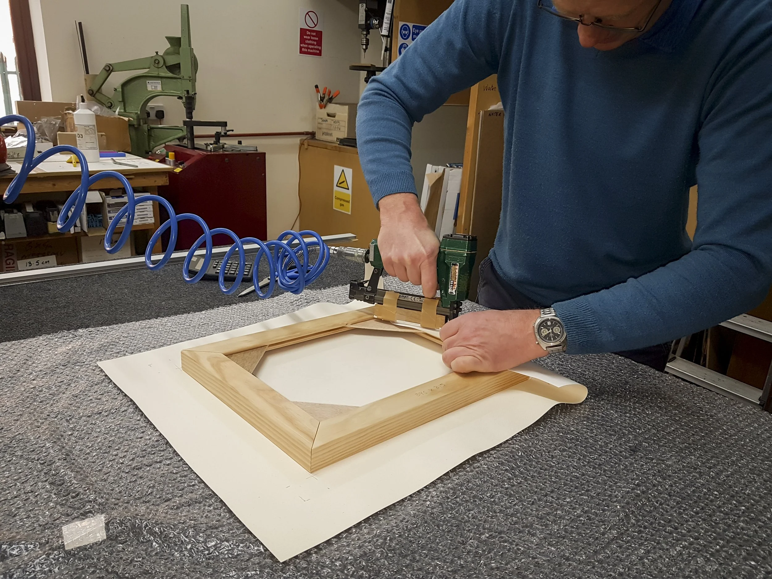 A person working on a woodworking project, using a nail gun to attach a wooden frame. The workspace is cluttered with tools, a workbench, and signage including safety warnings on the wall.