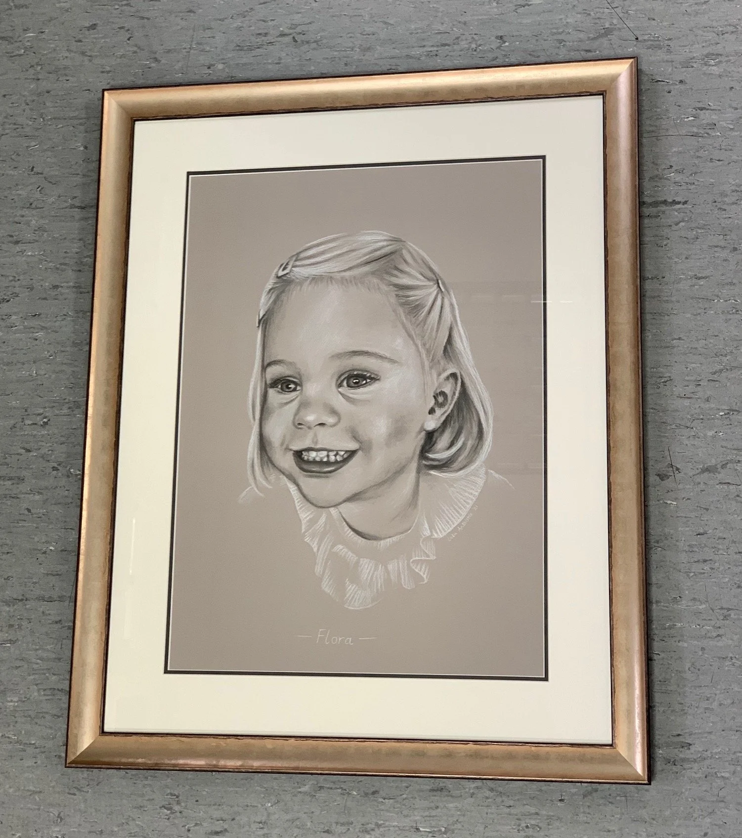 Black and white pencil drawing of a young girl with a big smile, short hair, wearing a ruffled collar with the name 'Flora' written beneath the portrait, framed and mounted on a gray textured wall.