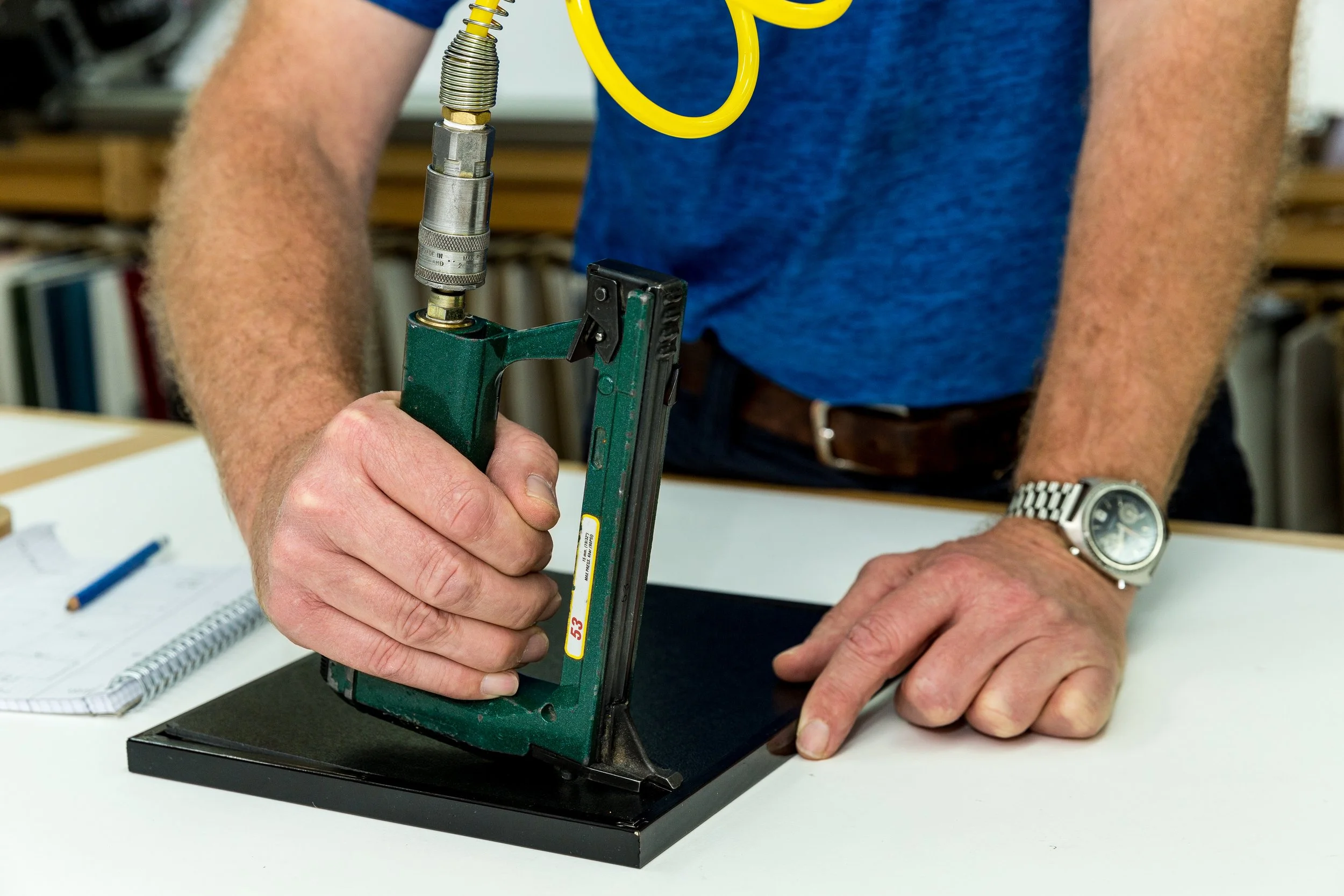 Person operating a mechanical press or crimping tool on a small solar panel or electronic component, with notebooks and pens on the table.