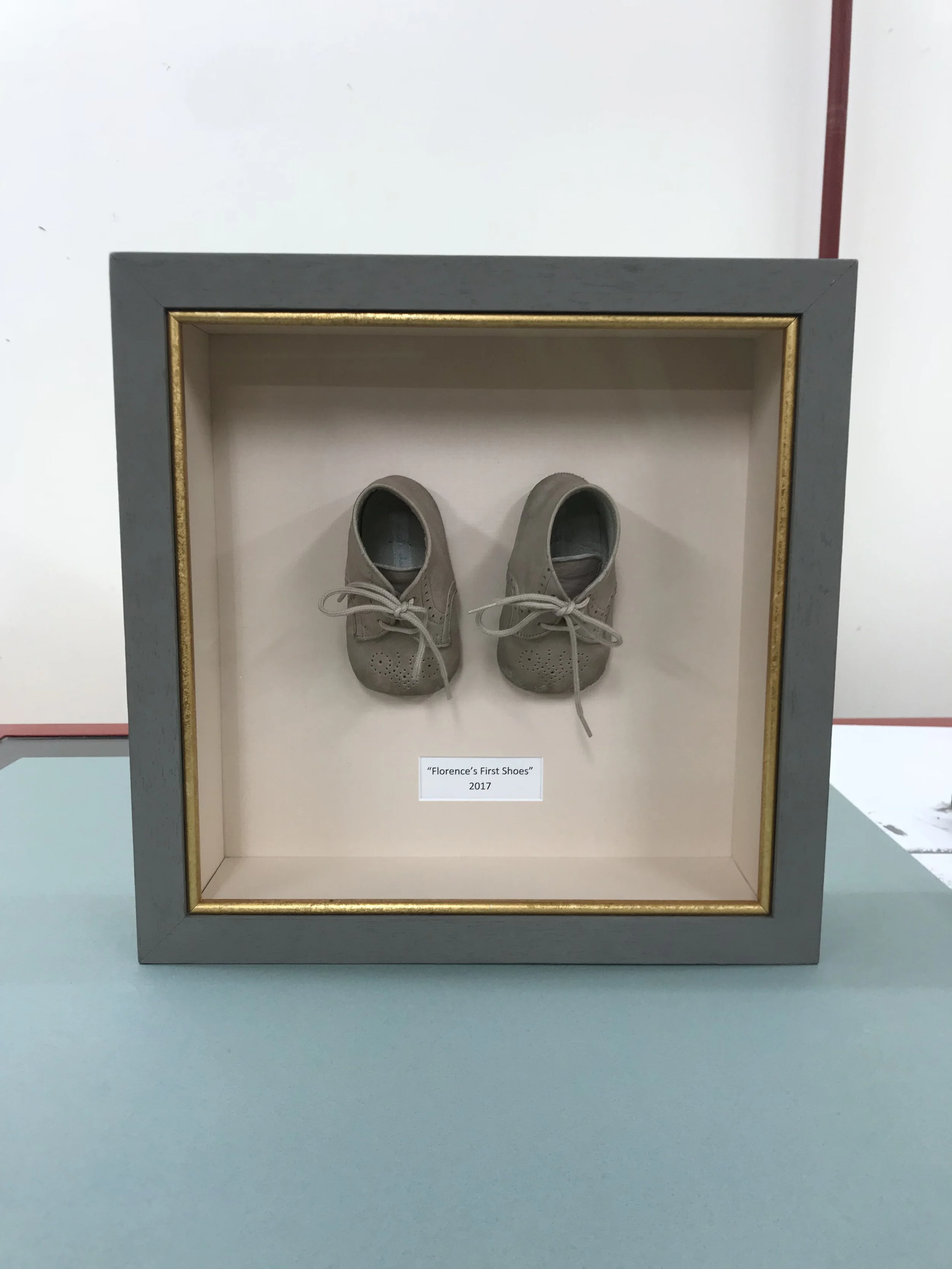 Display case containing a small pair of beige children's shoes with laces, labeled 'Florence's First Shoes' 2017.