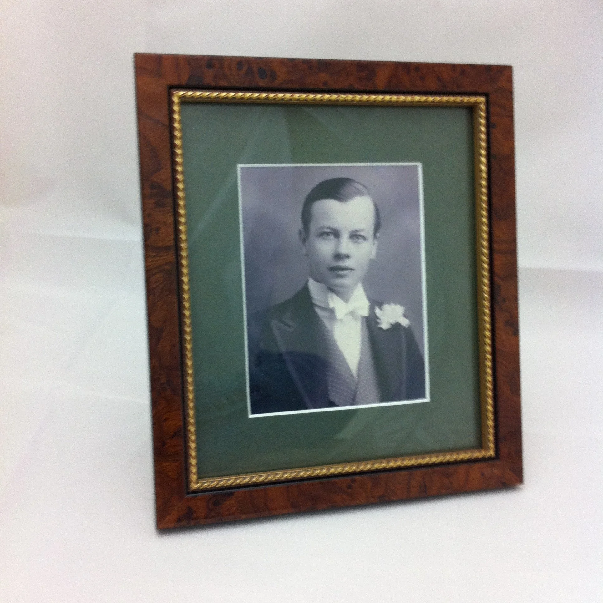 A framed black-and-white photograph of a man with slicked-back hair, wearing a tuxedo with a bow tie, and holding a white flower, hangs on a plain white wall.