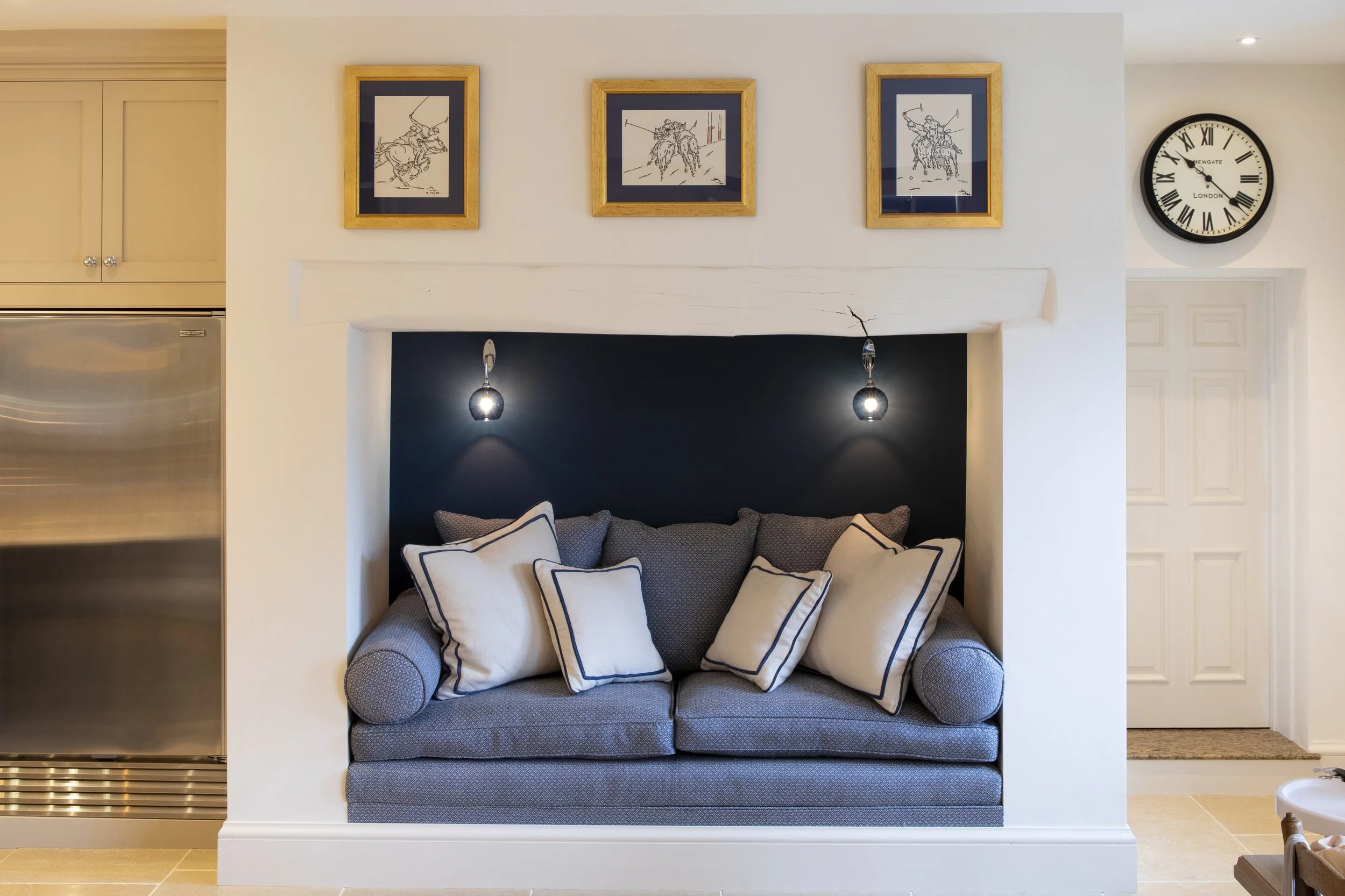 A living room with a gray couch with white and black throw pillows, a black wall behind the couch with two pendant lights hanging above, three framed sketches of horse riders on the wall, a round clock on the wall to the right, a white wall and doorw