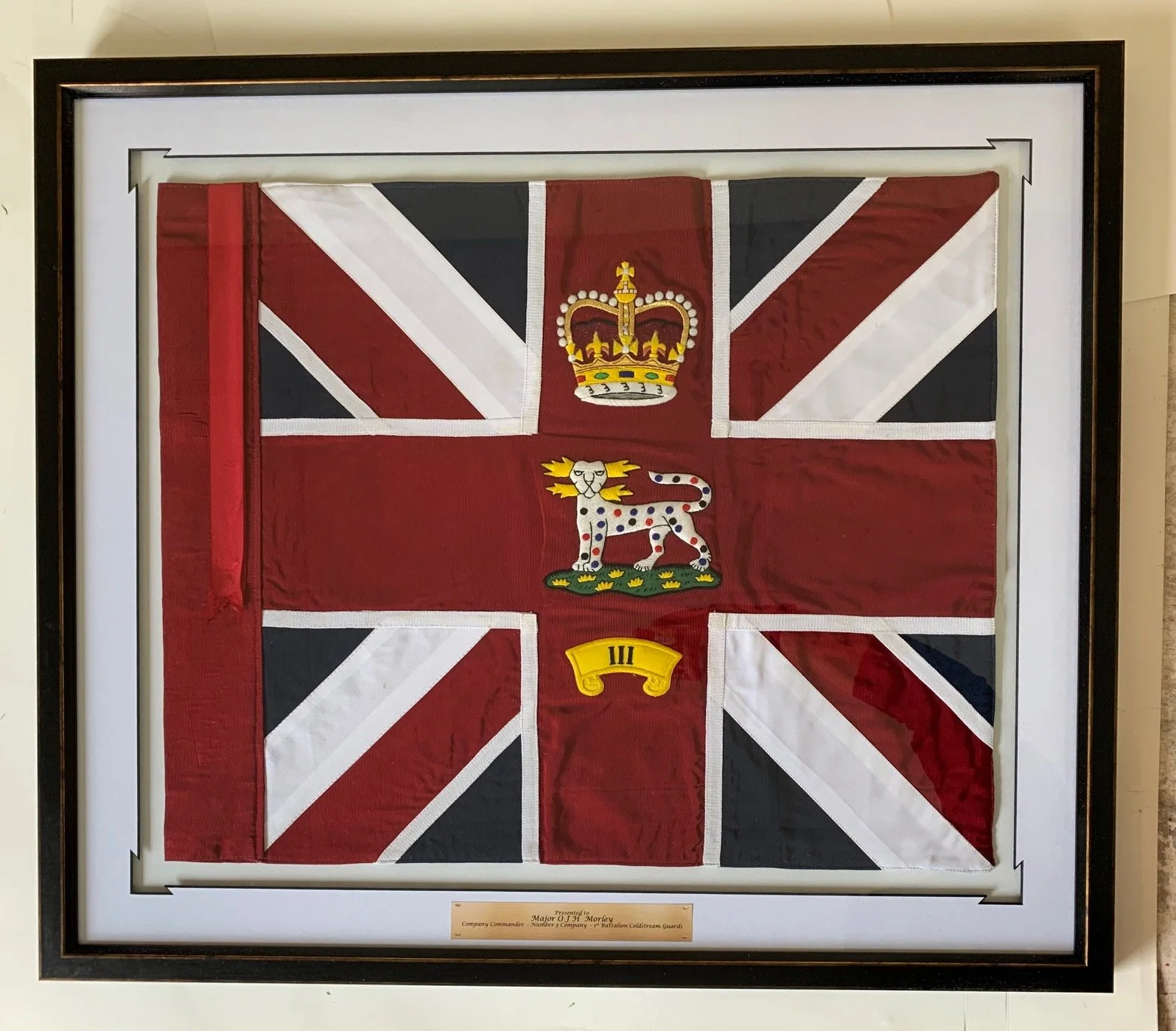 Framed display of a British flag with a crown, a white leopard with colored spots, and a yellow banner below it.