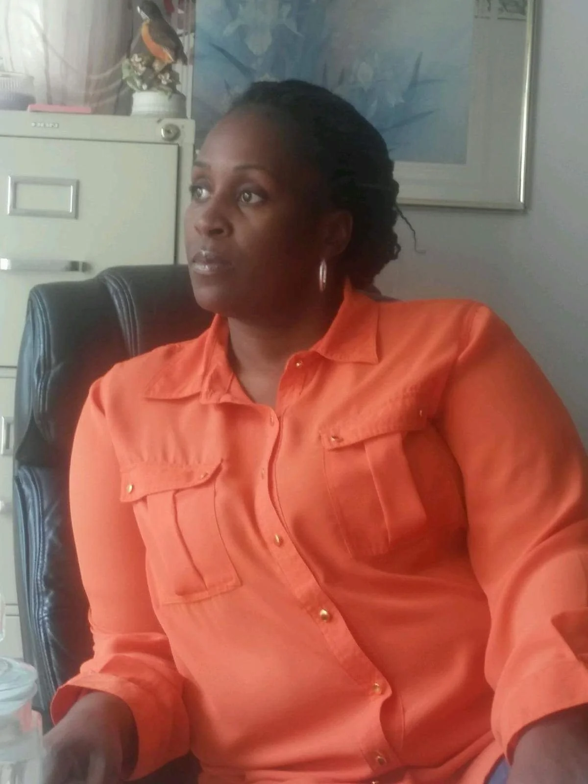 A woman with dark hair tied back, wearing an orange button-up shirt with pockets, sitting in a black office chair in an indoor setting with framed art and a filing cabinet in the background.