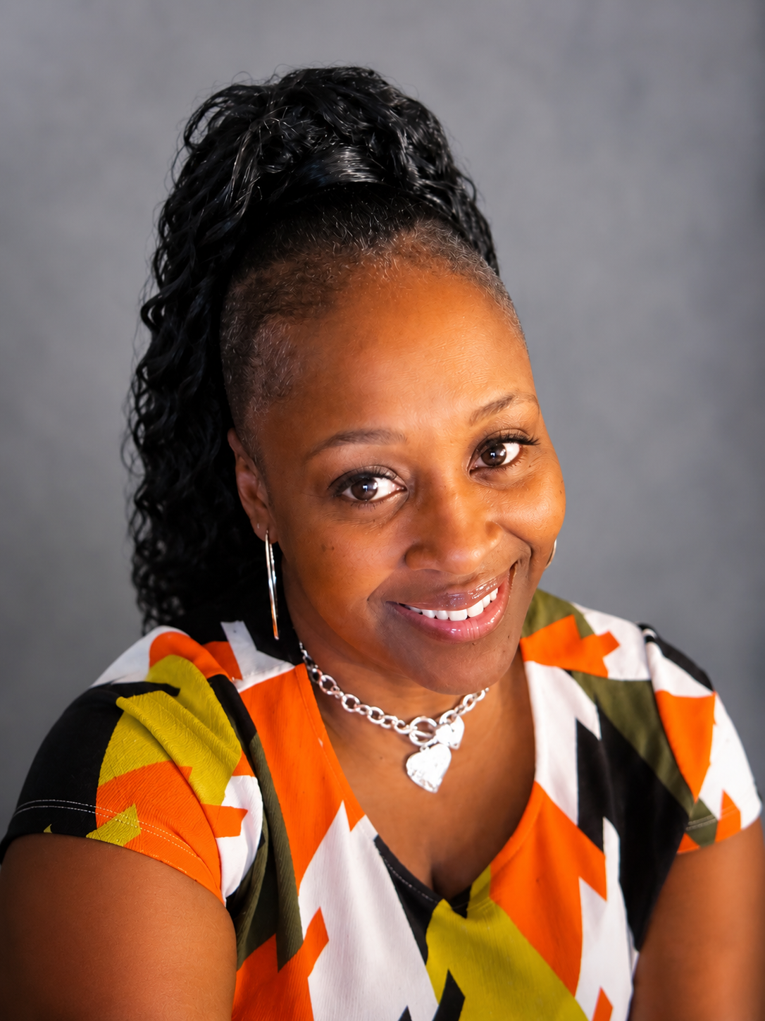 A woman with dark, curly hair styled in a high ponytail, smiling at the camera, wearing a multicolored patterned top, hoop earrings, and a heart-shaped pendant necklace against a plain gray background.