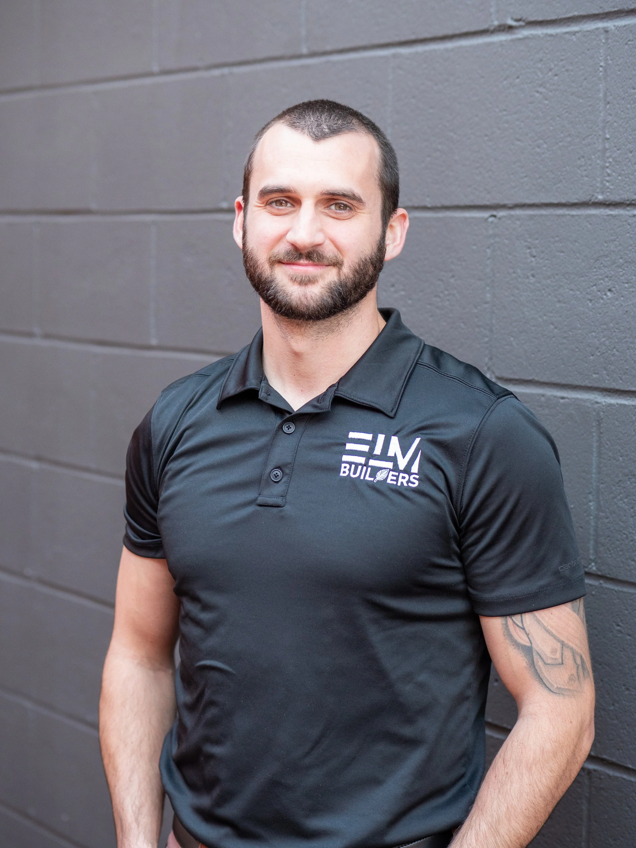 A man with a beard and short dark hair standing in front of a red brick wall, wearing a short-sleeved, button-up shirt with a small black and white checkered pattern.